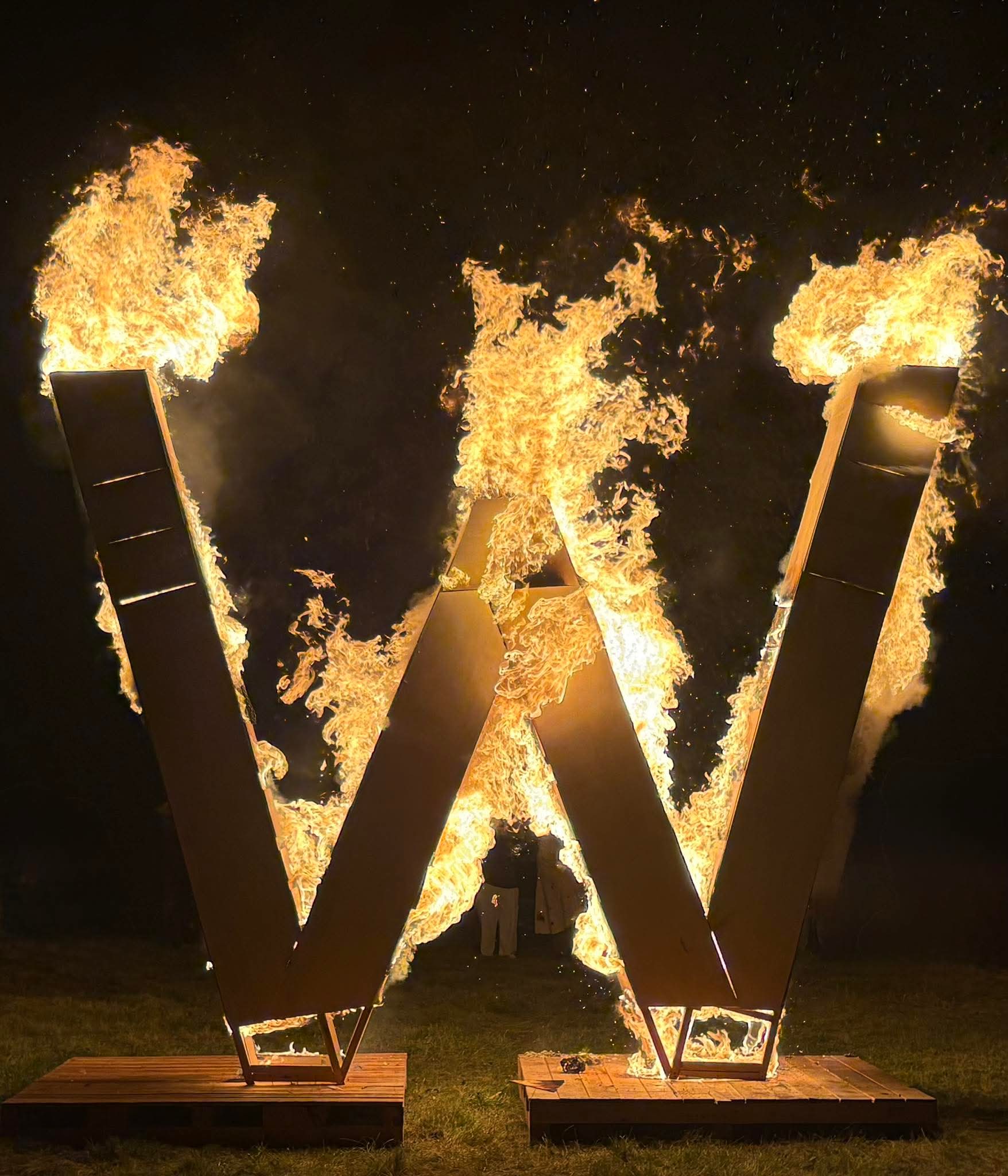 Two large, rectangular structures form a letter 'W' and are engulfed in flames at night.