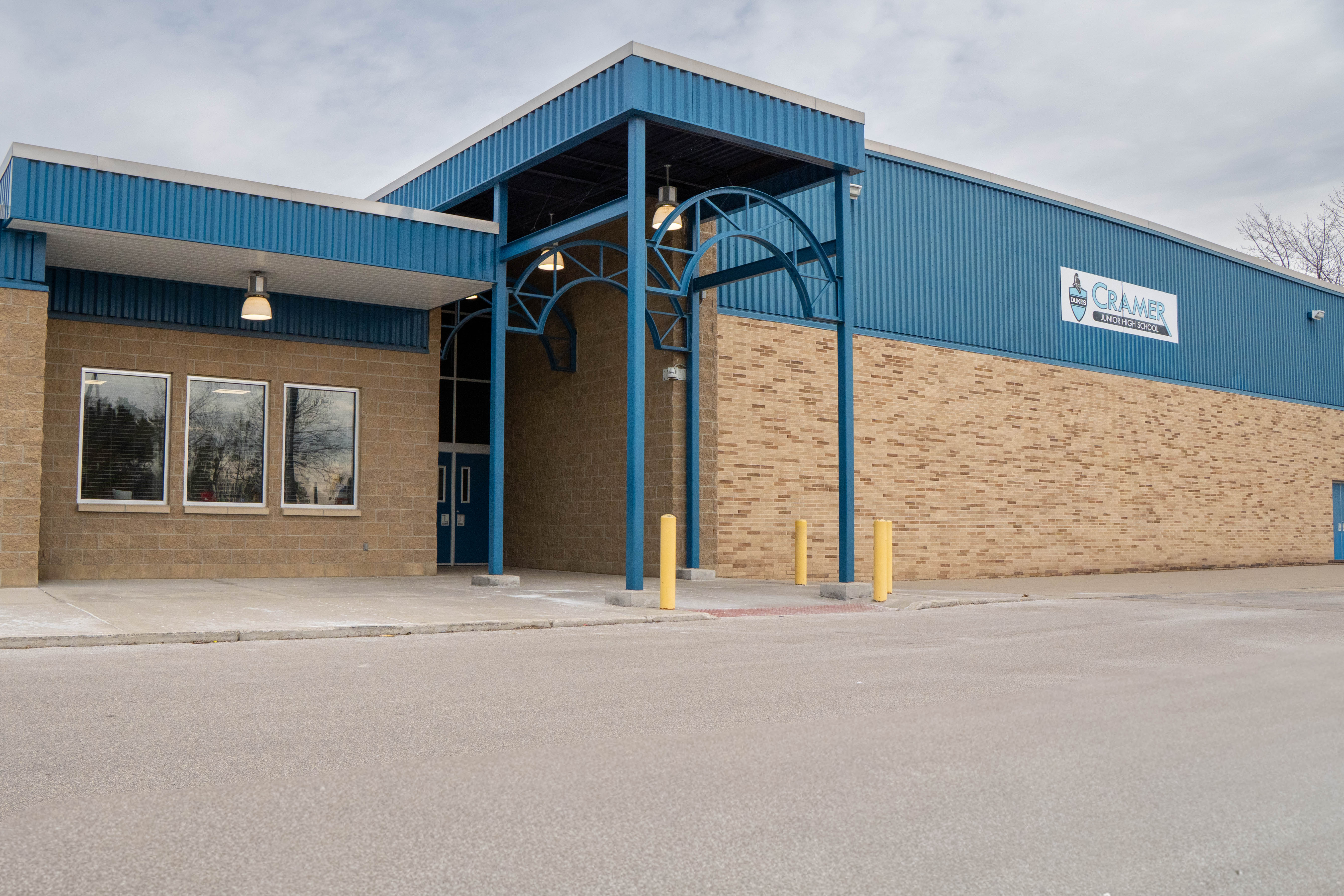 Front entrance of Cramer Junior High School featuring blue metal siding, tan brick walls, and a covered entryway with blue steel arches and yellow safety bollards.