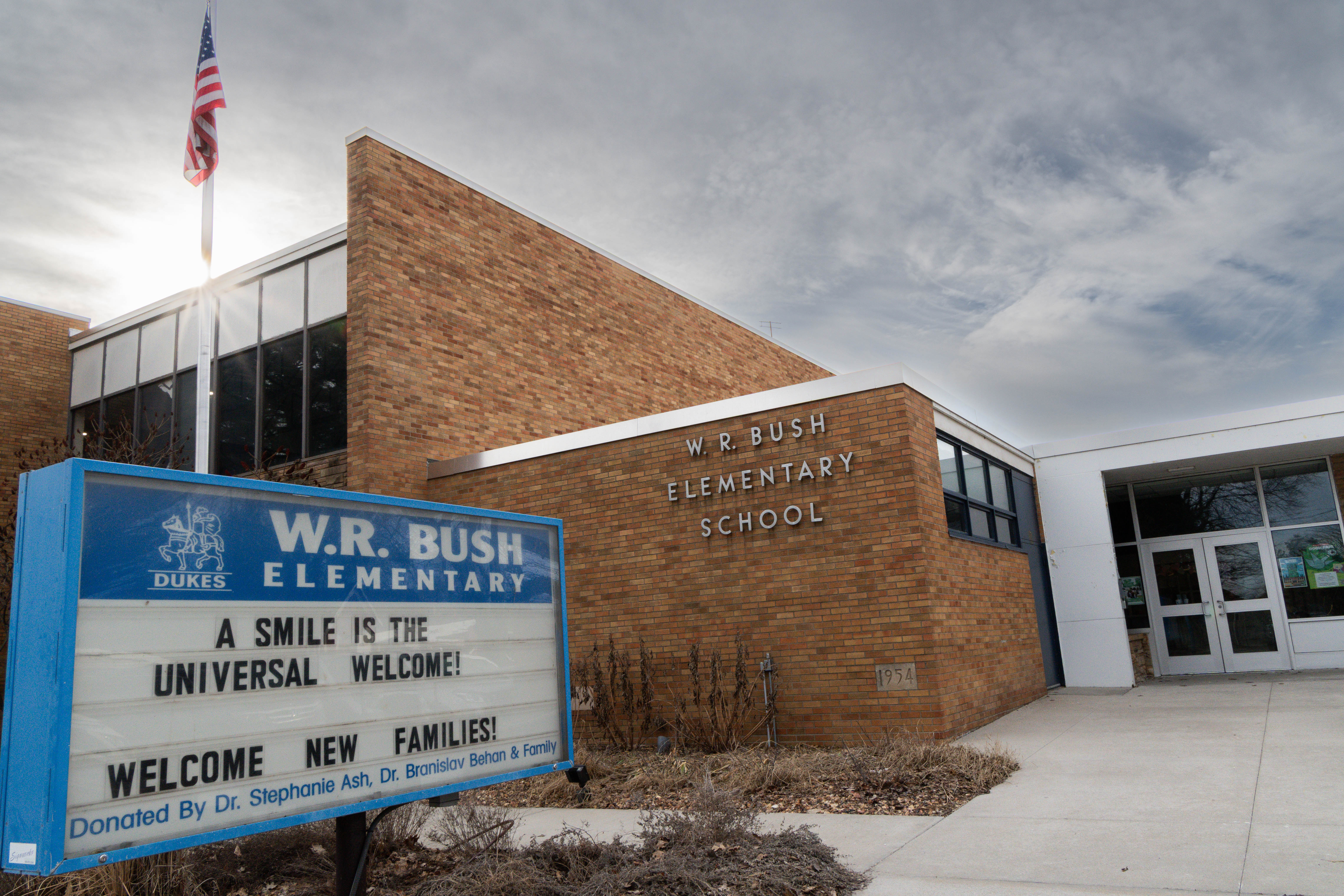 Exterior of W.R. Bush Elementary School with brick building, American flag, and welcome sign reading, “A Smile Is the Universal Welcome.”