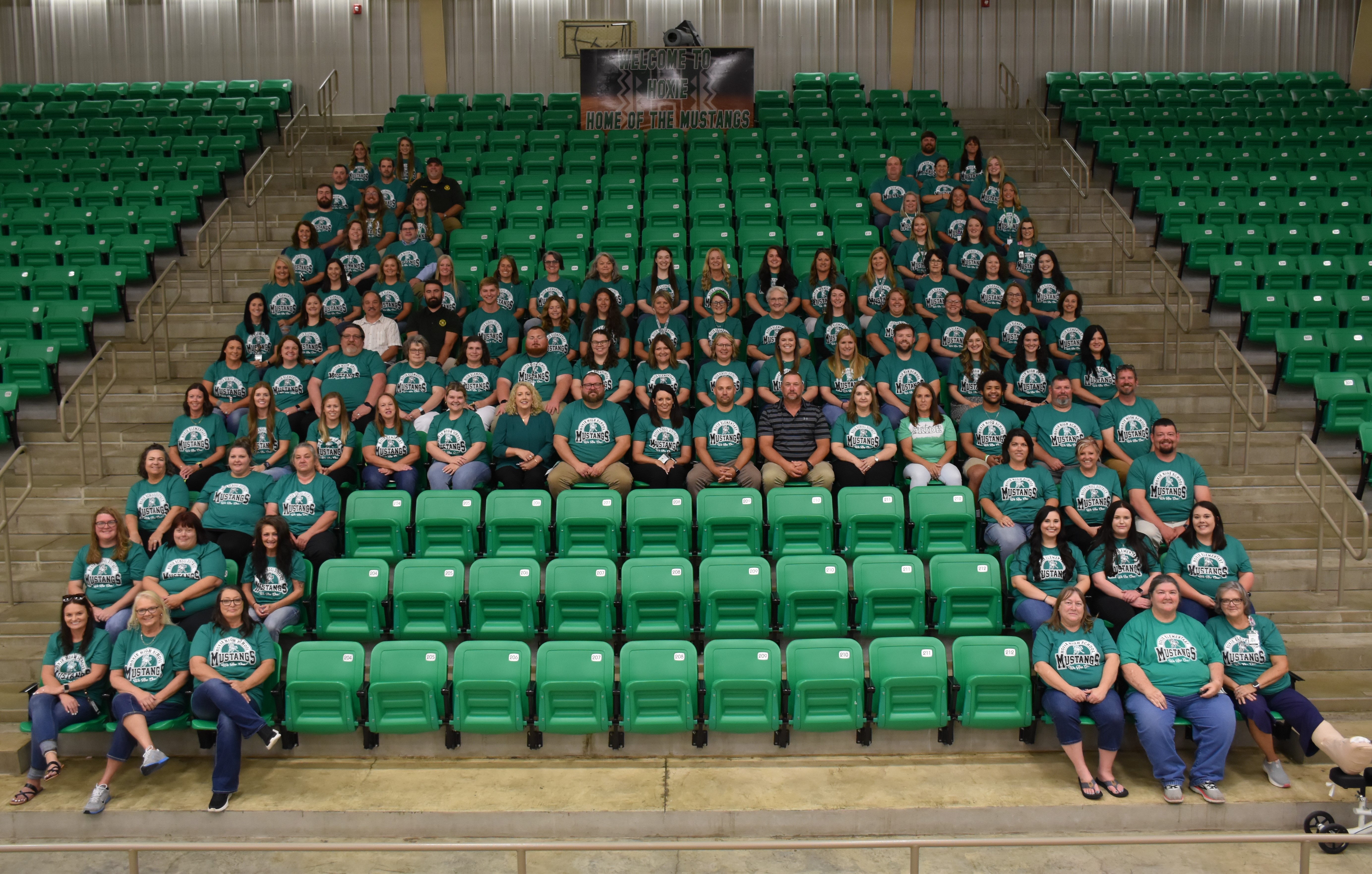 Employees in green shirts form a capital "H" by siting in bleachers.