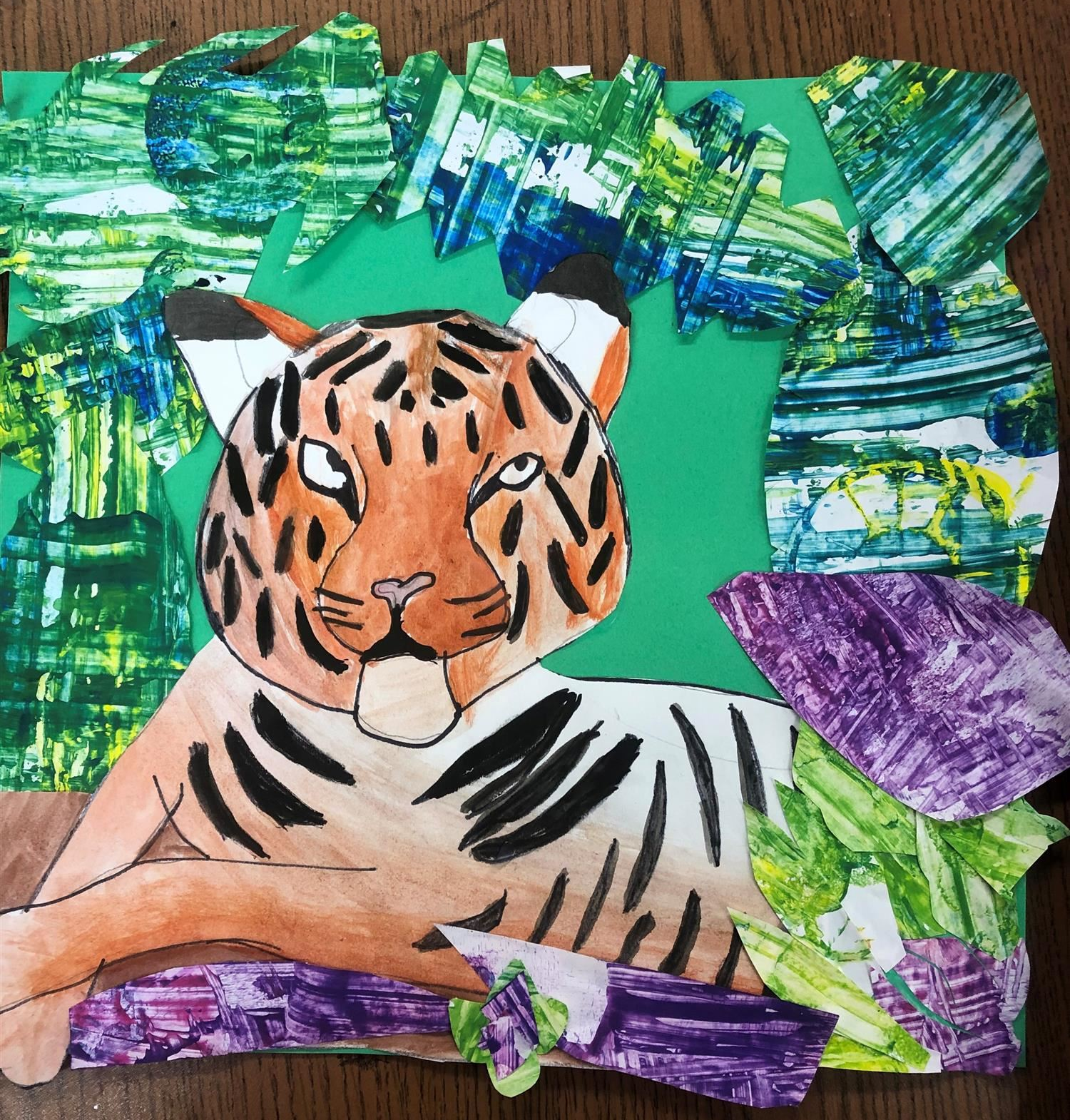 Drawing of an orange tiger laying in a field of paper cutout leaves. The leaves are painted green, blue and purple.