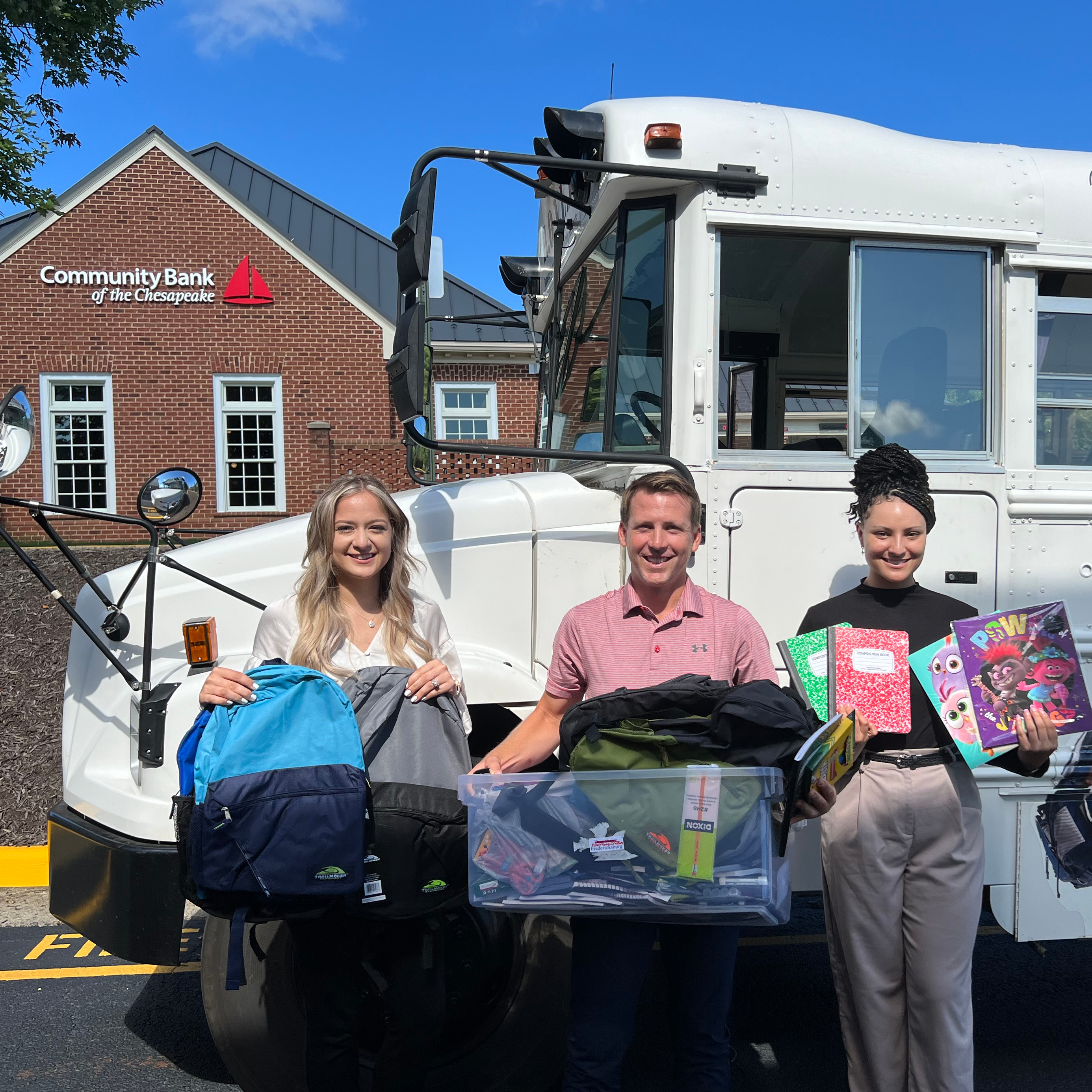 A group of people smiling and holding up school supplies in front of the white rover school bus. They are from the community bank of the chesapeake.