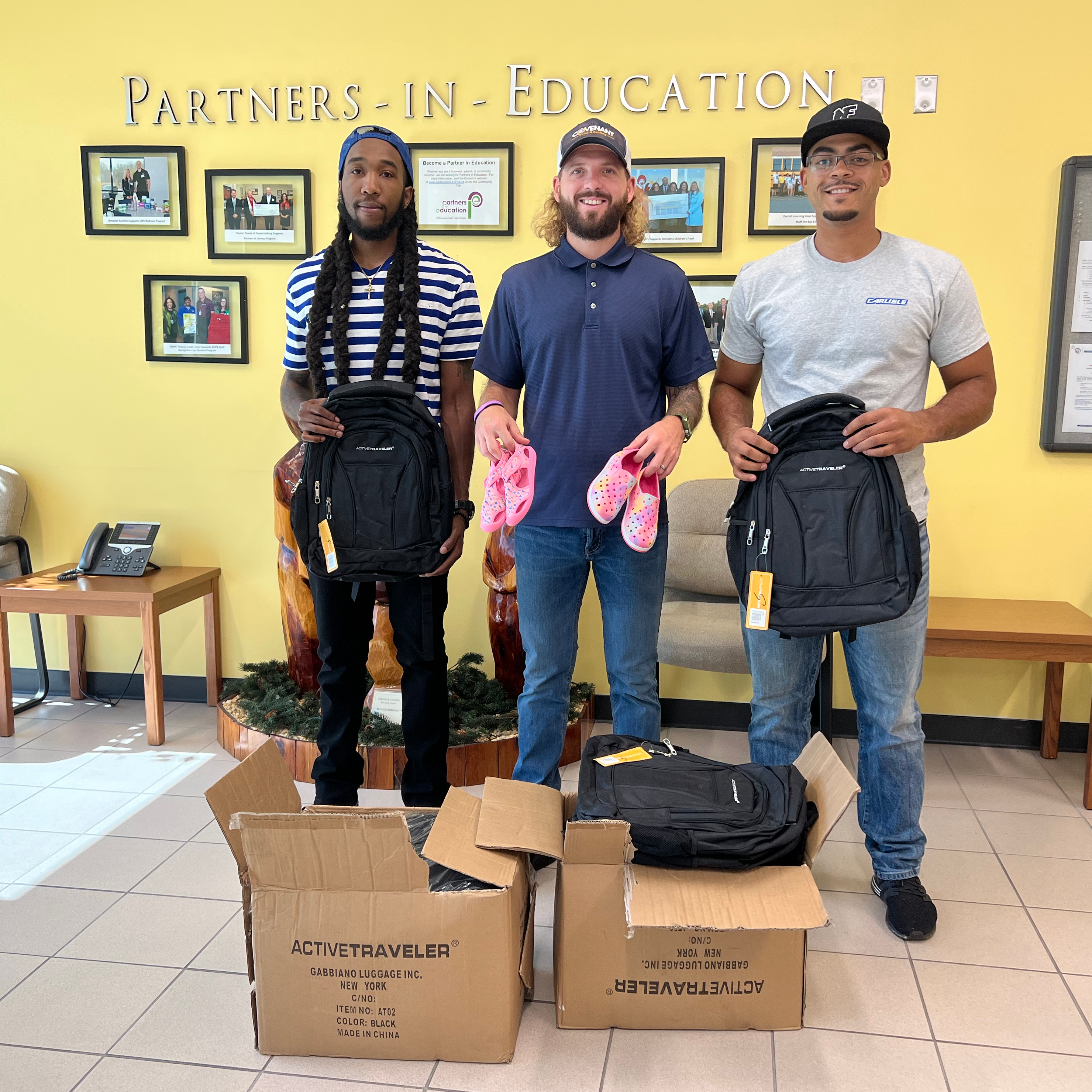 Three men from Kicks for Kids pose with the shoes and backpacks they donated in front of a yellow wall reading "Partners in Education".