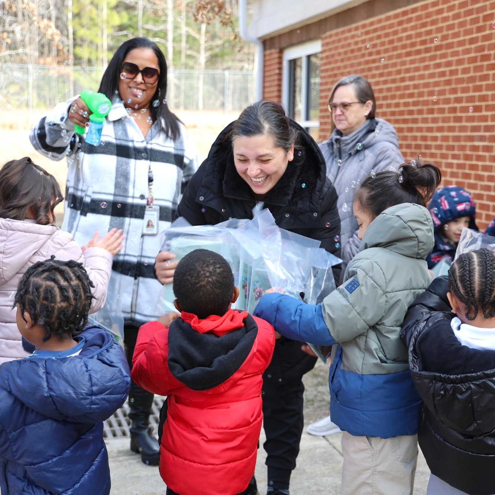 Adults and children gathered outside a brick building. Adults distribute plastic bags to children, who hold them.