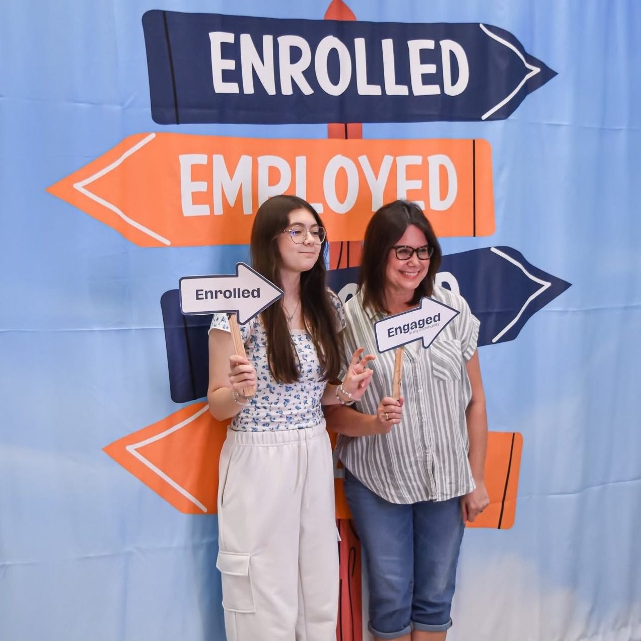 Two women stand in front of a banner with "Enrolled" and "Employed" arrows, holding signs reading "Enrolled".
