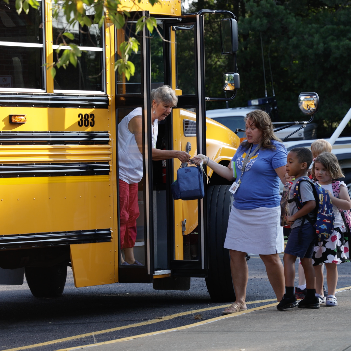 People in front of a yellow school bus, including a woman and a child, on a sunny day.