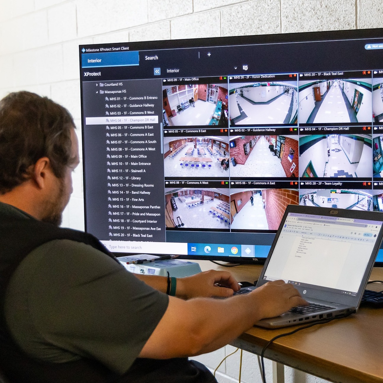 Person in front of a computer monitor and laptop, viewing multiple video screens with indoor views.