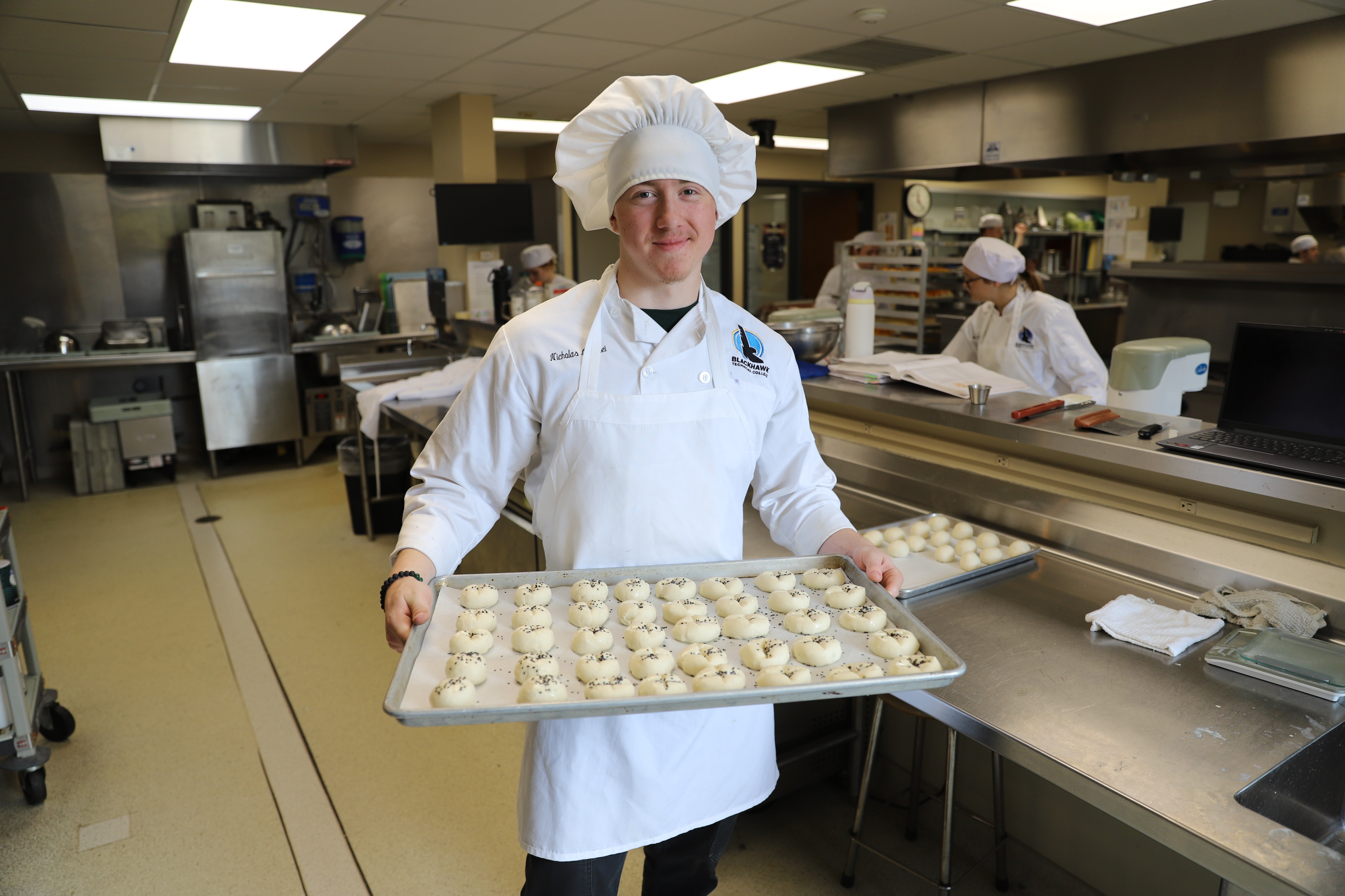 A person in a chef's uniform and hat holds a tray of unbaked pastries in a kitchen.