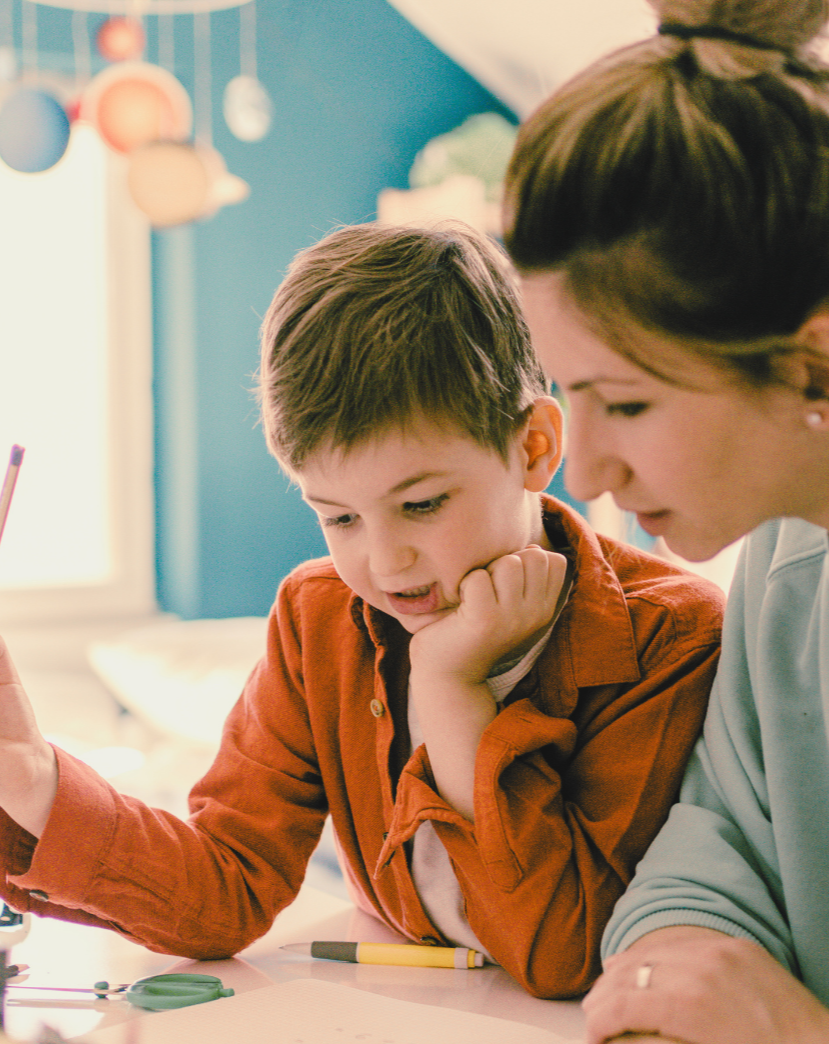 Adult and child sitting at table. Child holds pencil and looks at paper. Adult looks at child.
