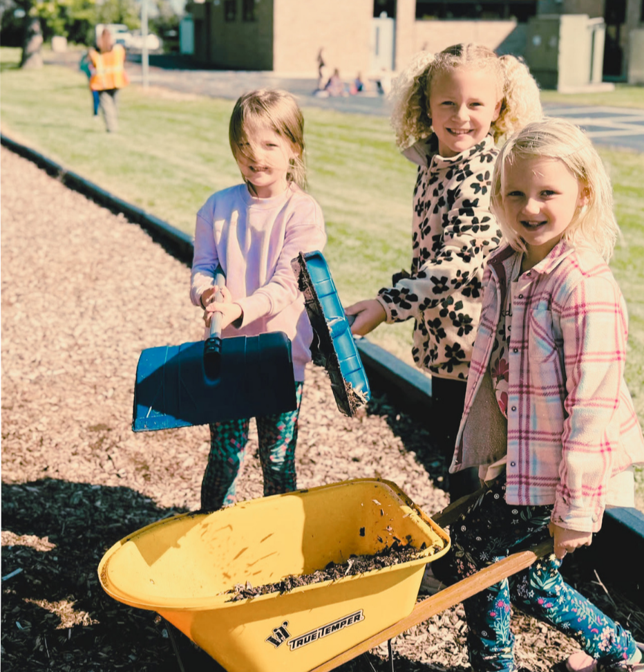 Three young girls, one holding a blue object, another pushing a yellow wheel barrow, and the third holding a blue item, all standing on a pathway.