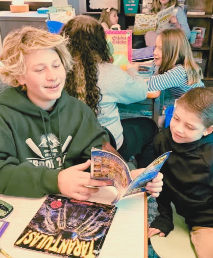 Kids sitting at a table with one reading a book aloud to another. Other students and teachers in the background.