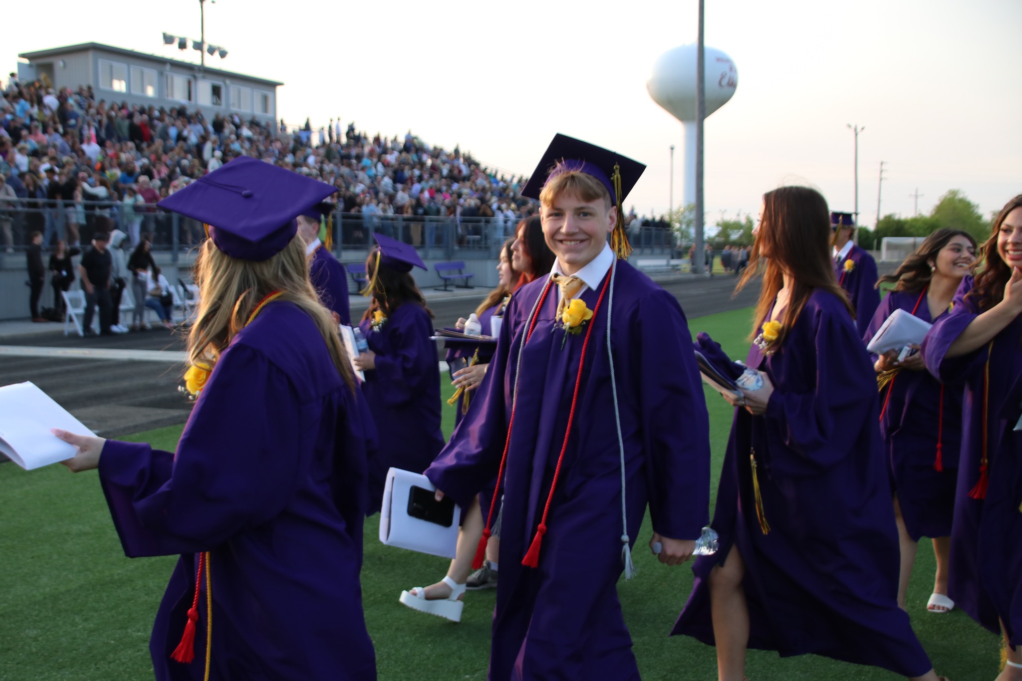 Graduates in purple gowns and caps walk across the field while holding papers; spectators are seated in the background.