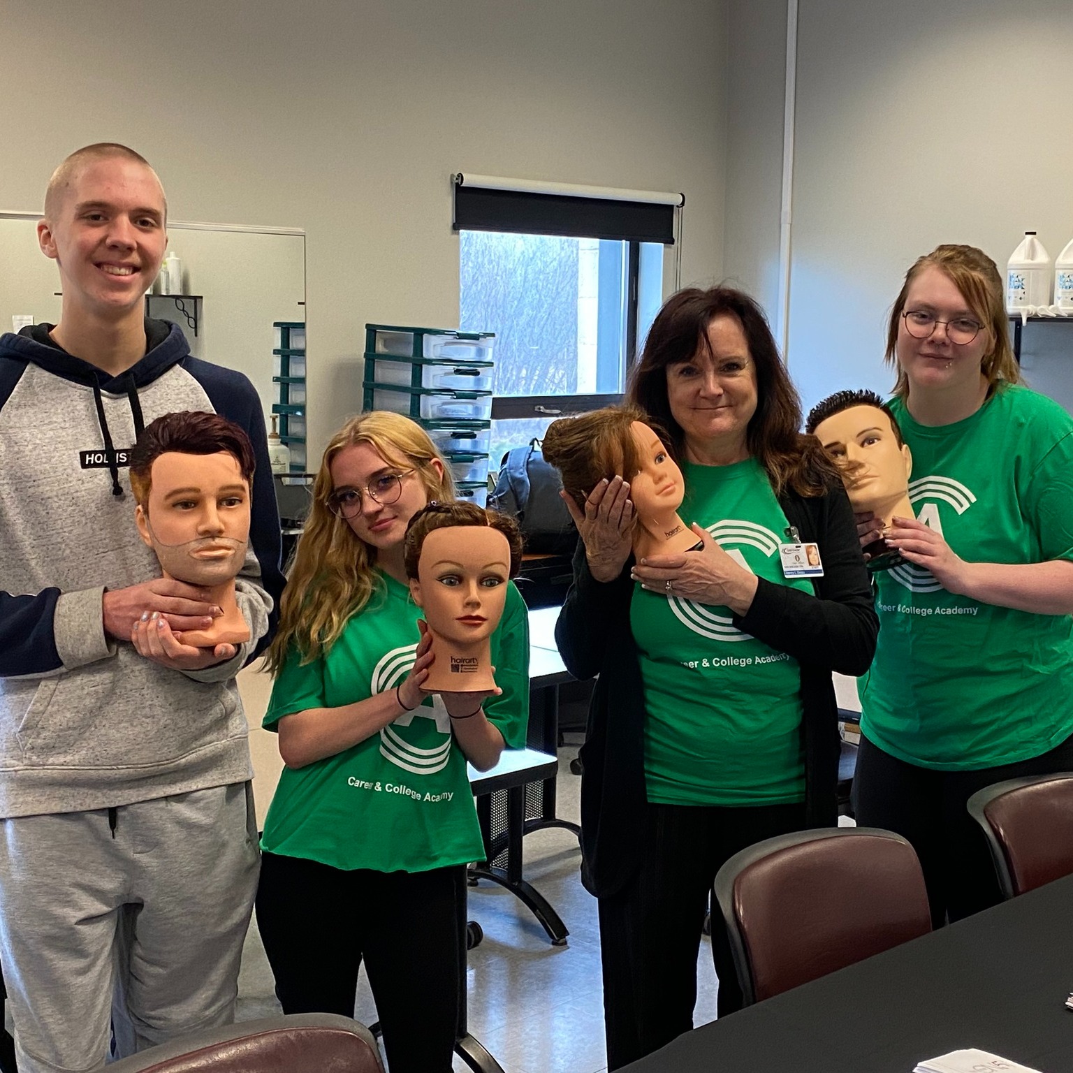 A group of people in green shirts stand in a classroom holding mannequin heads. They are posing for a photo.