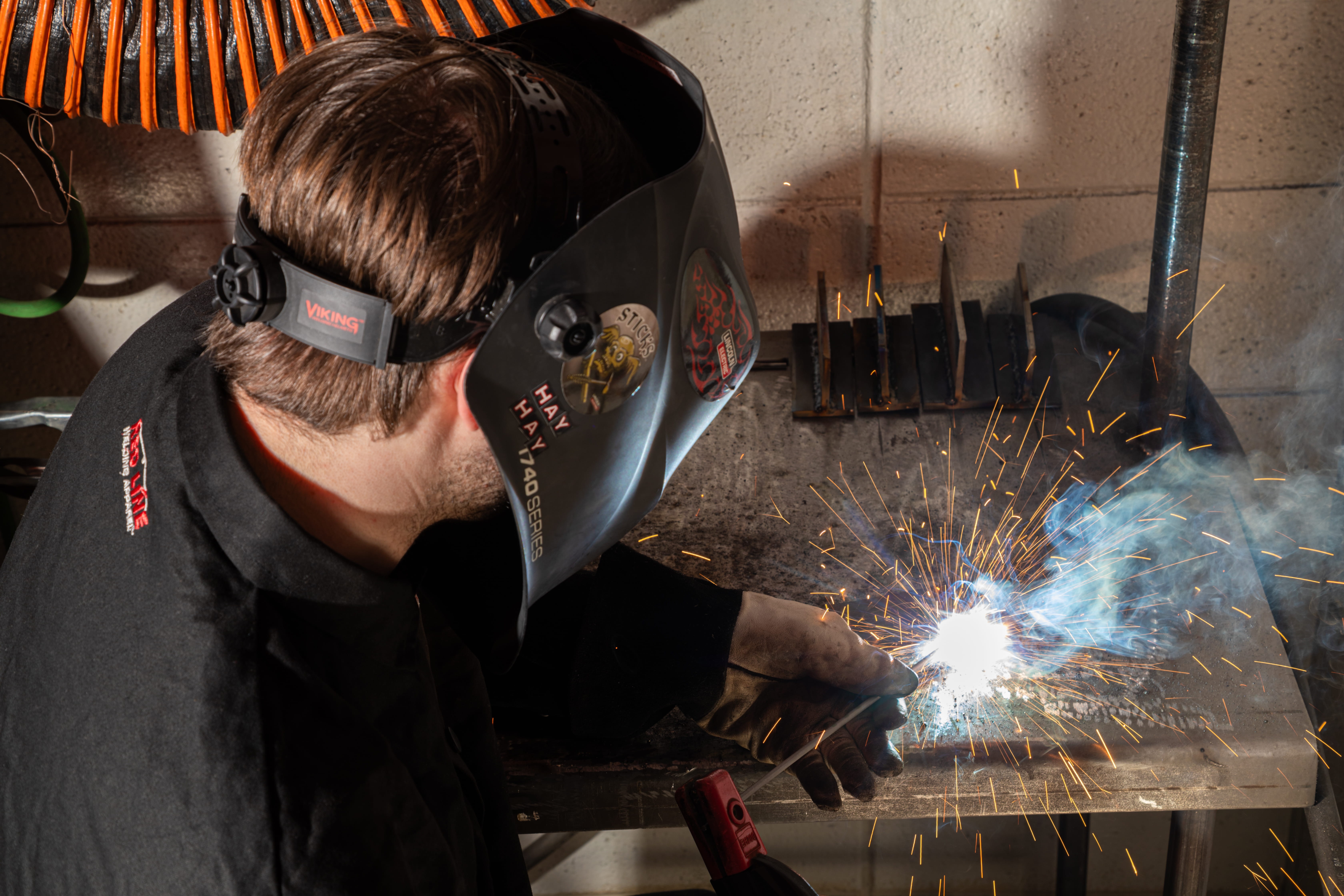 A person wearing a welding helmet and black shirt is welding metal in a workshop. Sparks are flying.