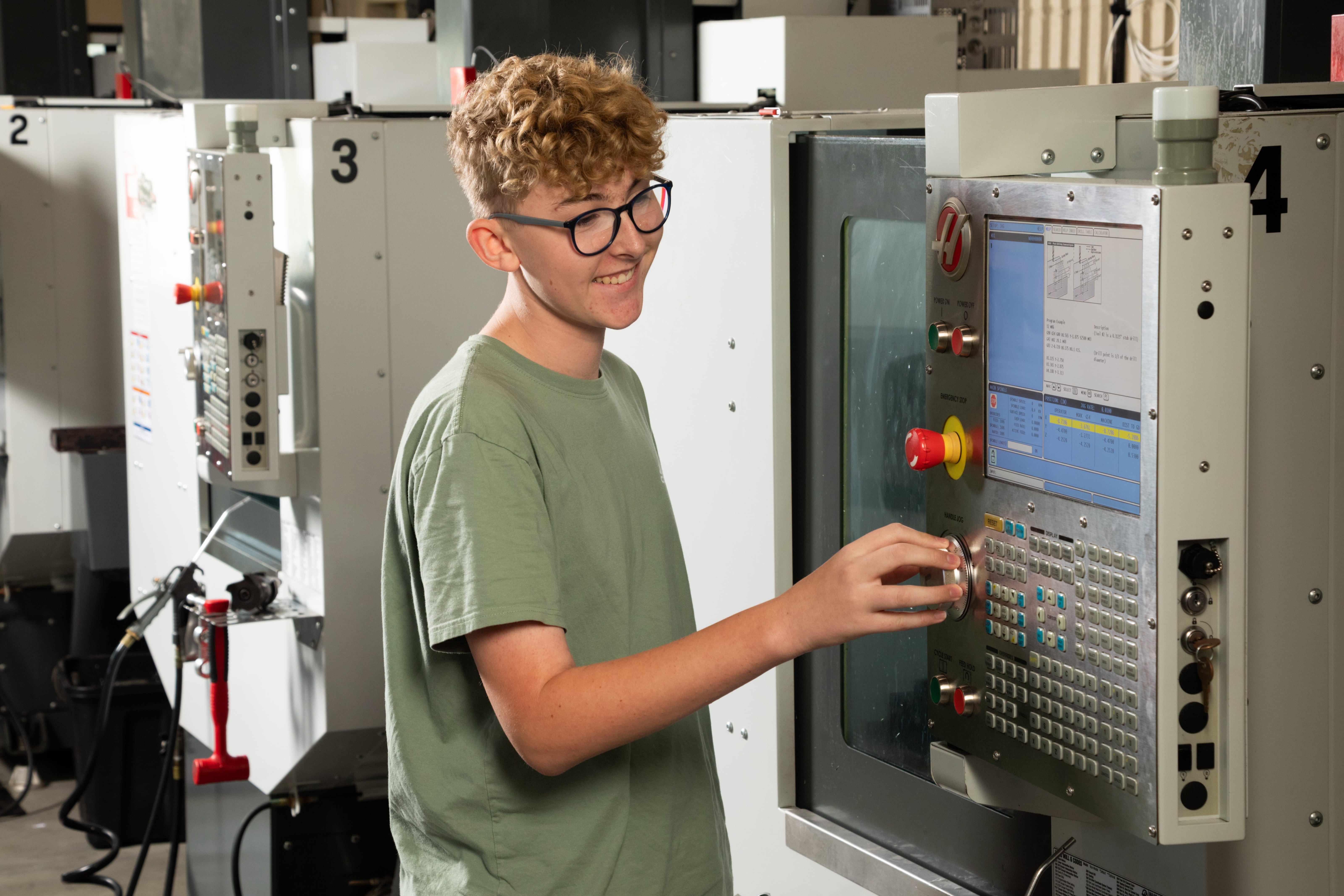 A person wearing a green shirt and glasses operates a machine with a touchscreen display in a factory.