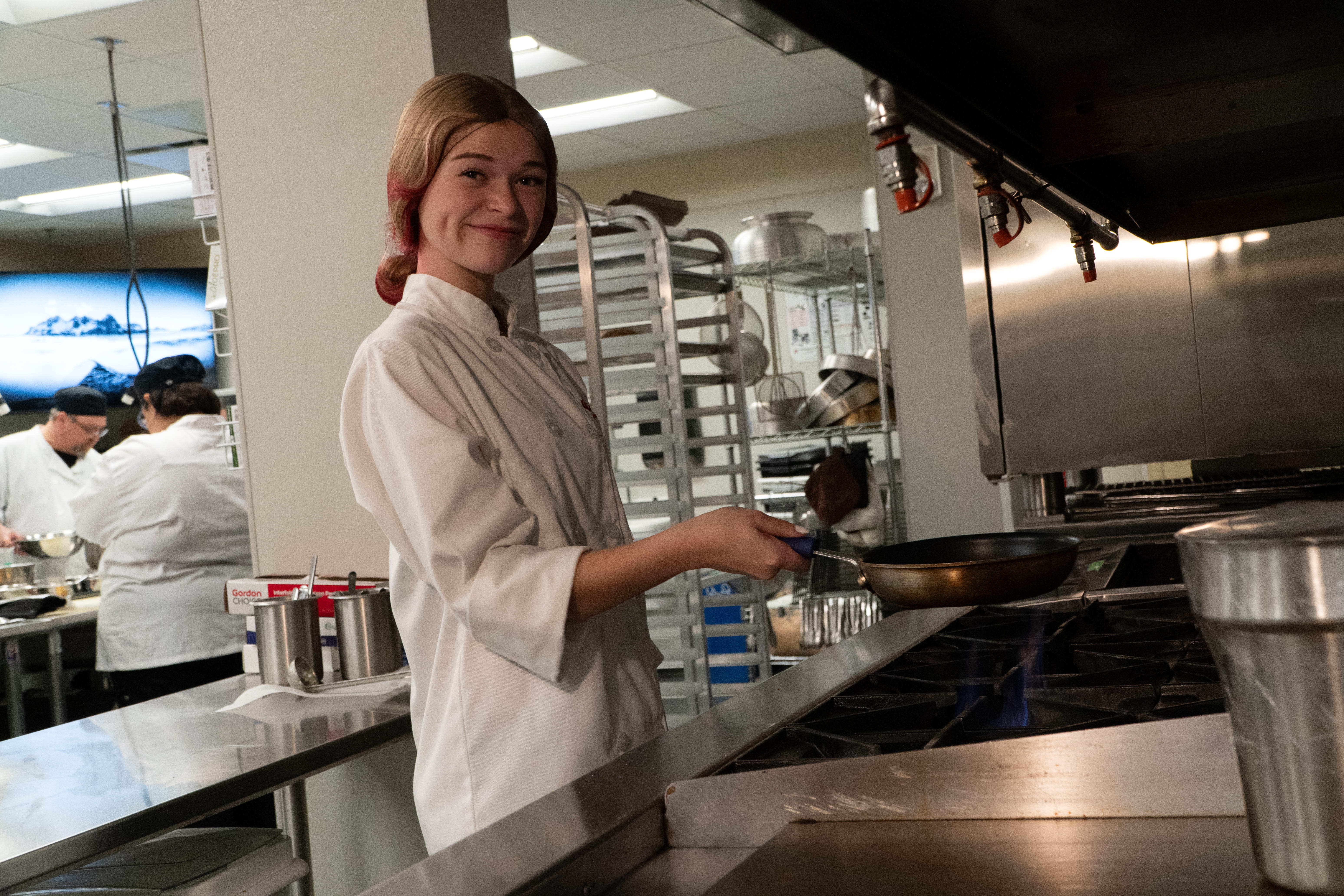A woman in a white chef's uniform stands in a kitchen, holding a frying pan above a stove.
