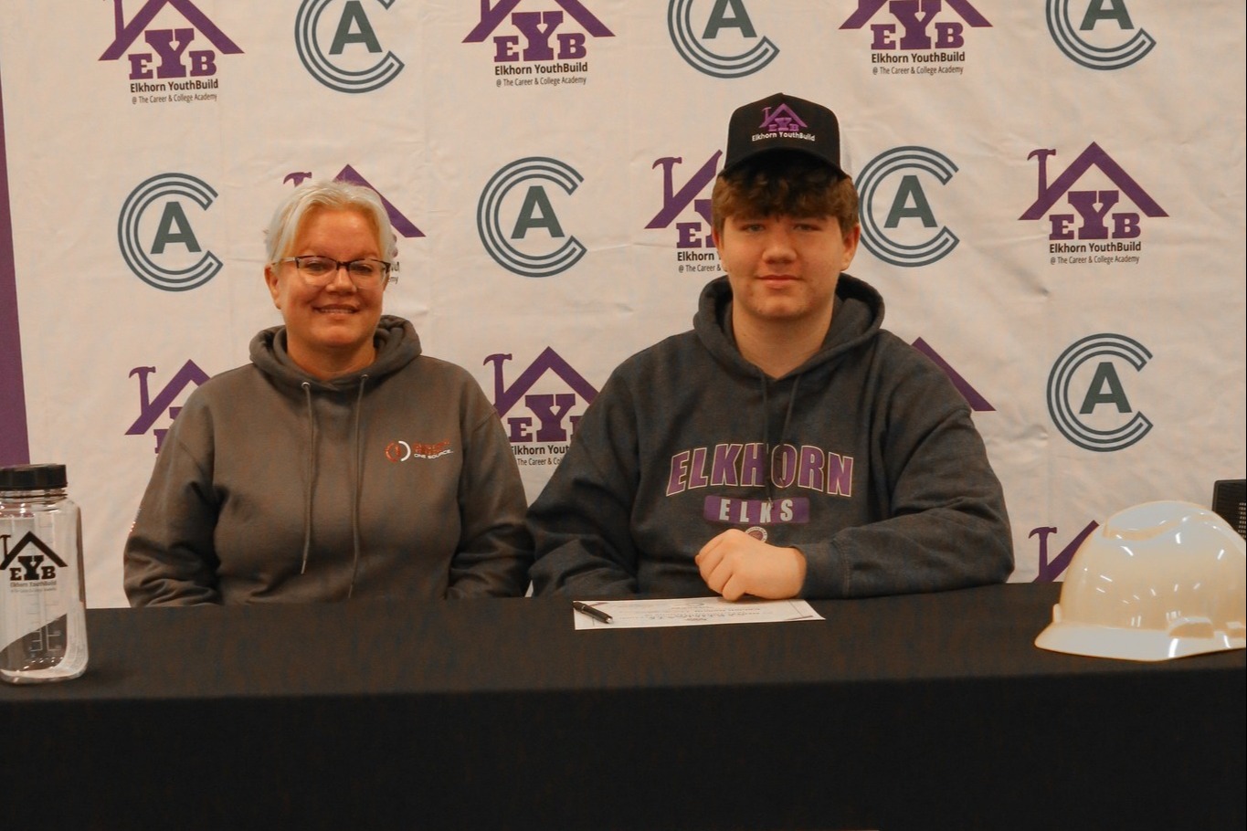 Two individuals sit at a table with a banner behind them. The banner reads "Career & College Academy" and has logos.
