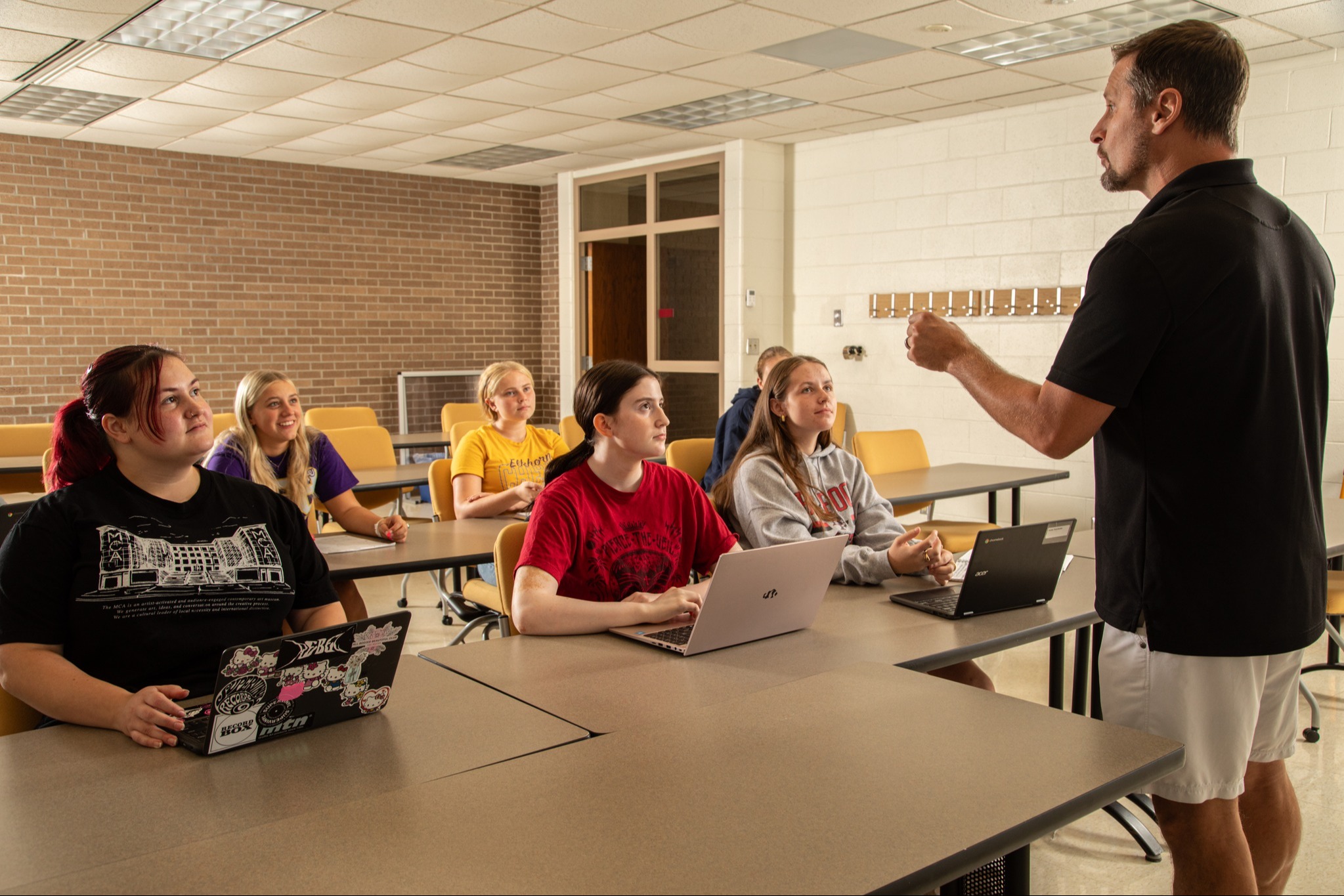 A man stands in front of students in a classroom, speaking. Students sit at tables with laptops.