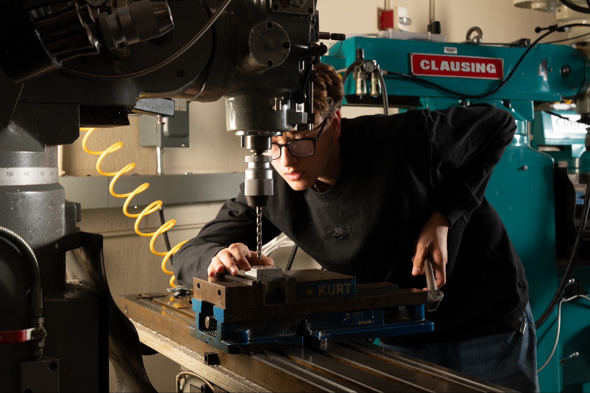 A person wearing glasses and a black shirt operates a machine in a workshop. Behind them is a green machine.