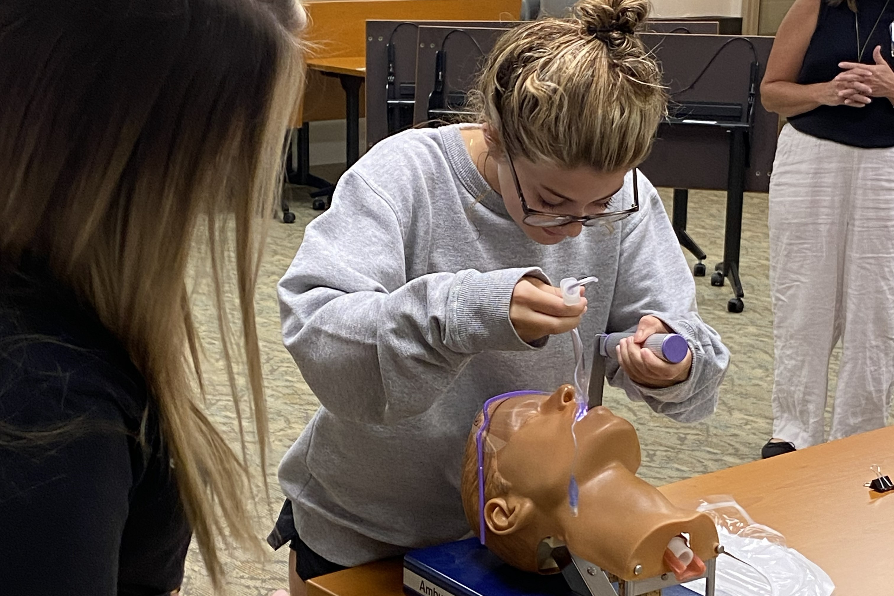 Two women in a room with a mannequin on a table. One woman uses a tool on the mannequin's face.