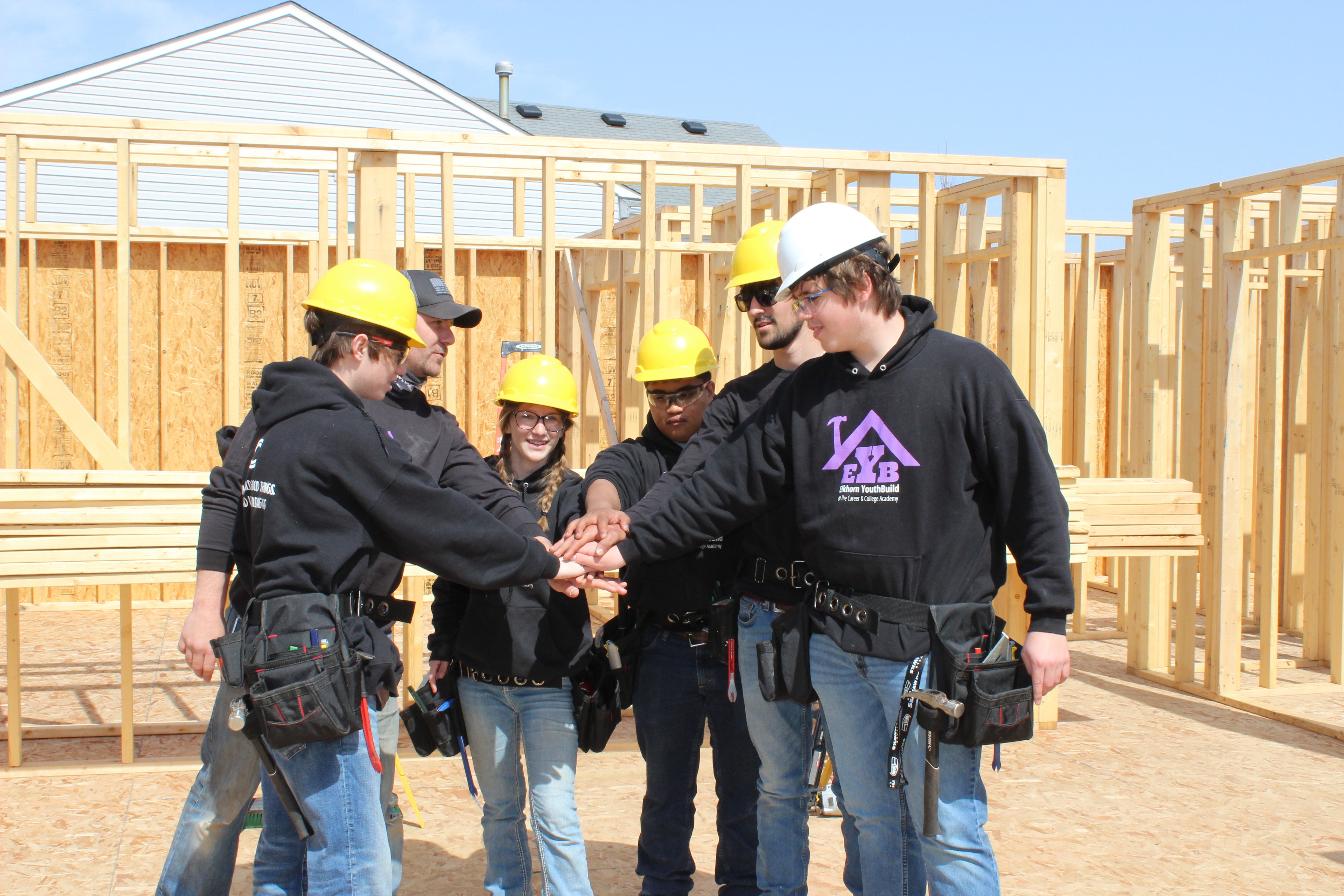 Workers in hard hats shake hands in front of an unfinished house. The house is made of wood.