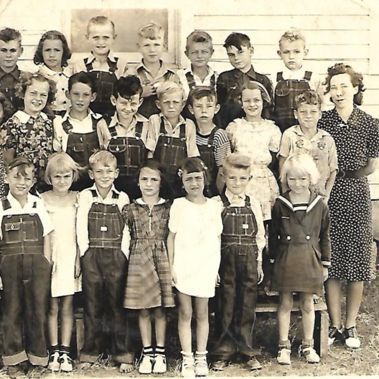 A black-and-white photo of a classroom with children in simple, vintage clothing and an adult standing near a door.