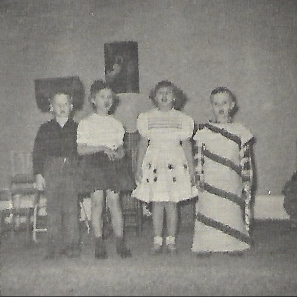 Four children in various clothing, one in a striped dress, stand on a stage. A picture frame hangs on the wall behind them.