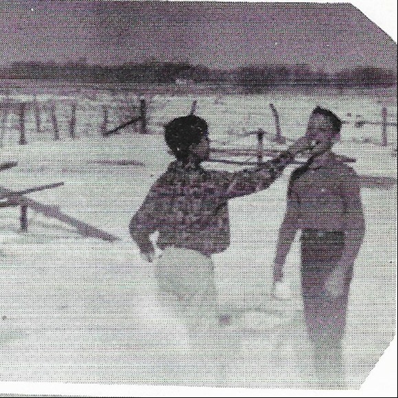 A black-and-white photo of two people standing in snow. One person holds the other's hand near the mouth. Behind them, there is a fence and snow-covered ground.