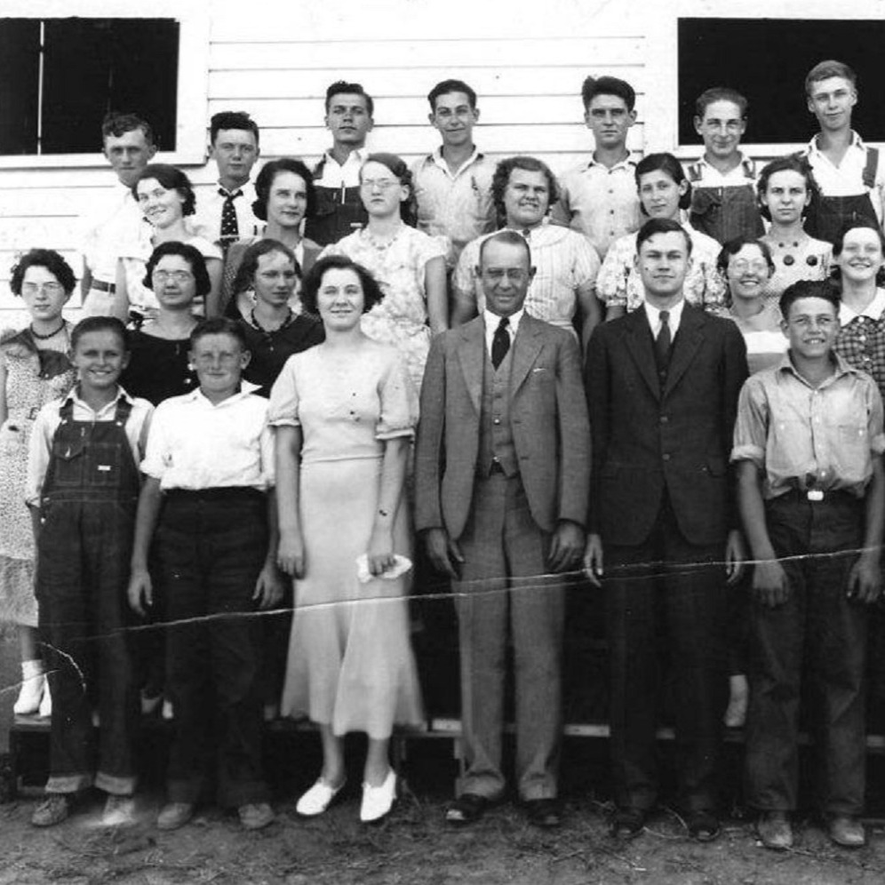 A group of people, including adults and children, stands in front of a building in a vintage black and white photo.