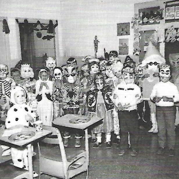 Black-and-white image of a classroom where children in Halloween costumes stand in a line.