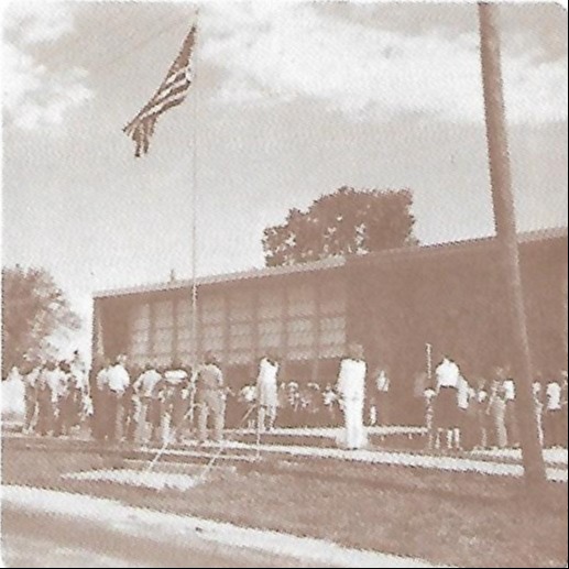 Monochrome photo of people standing in front of a building with an American flag on a pole.