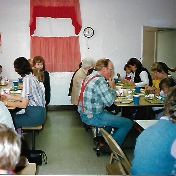 Several people sit around tables in a dining area with white walls, red curtains, and a clock.