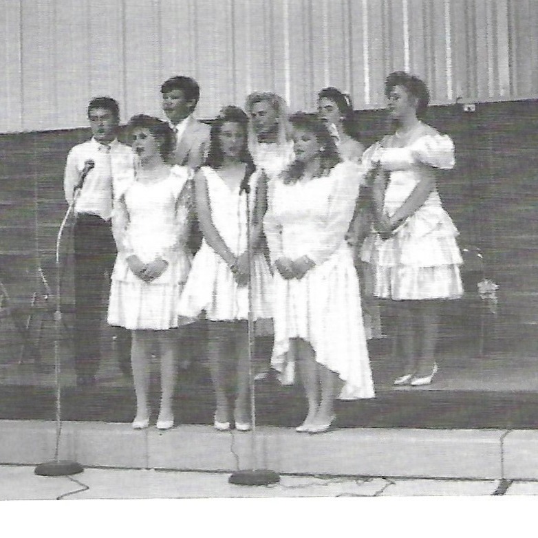 Black-and-white photo of a group of women in white dresses and men in suits on a stage with microphones.