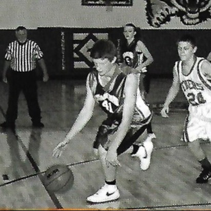 A black-and-white basketball game with players in jerseys, sneakers, and shorts on a wooden court. A referee stands on the sidelines.