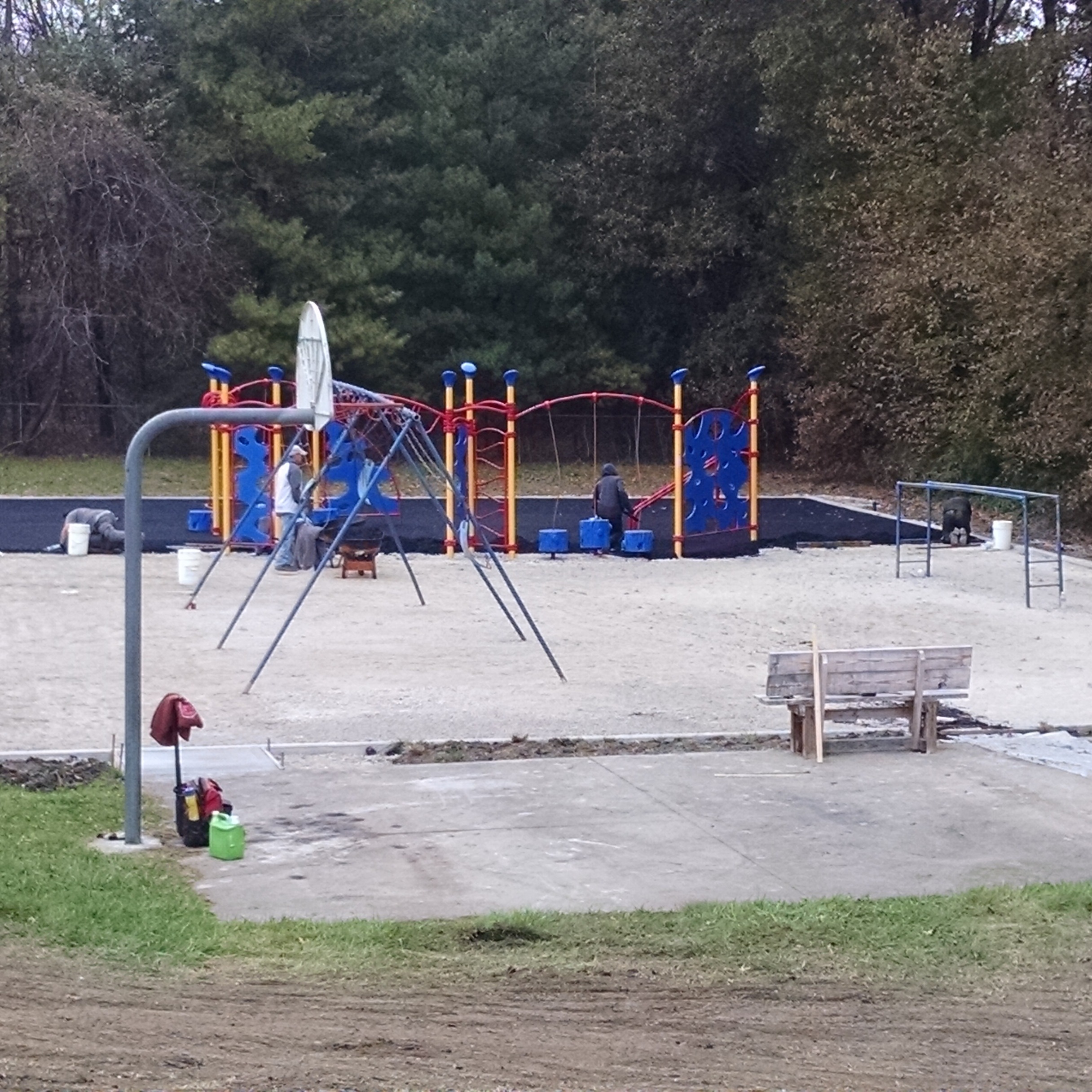 Playground with slides, swings, and benches on a dirt path surrounded by green grass and trees.