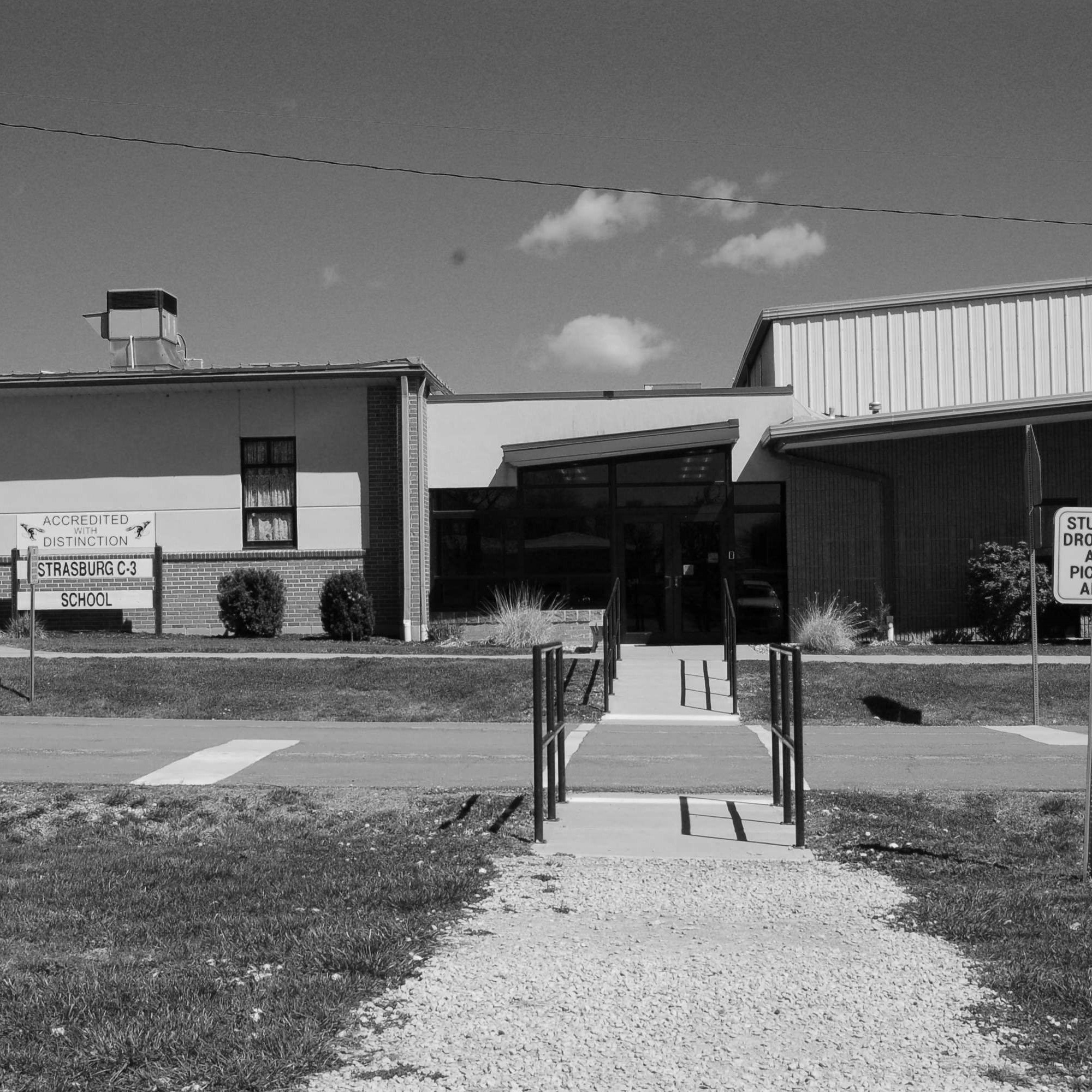 A black-and-white photo of a building with a ramp leading to its entrance. A sign on the sidewalk is visible.