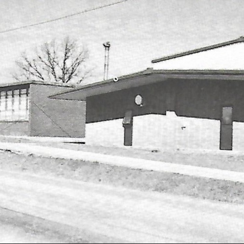A black-and-white photograph of a building with windows, a satellite dish, and a sidewalk. The building is adjacent to another structure with trees behind it.