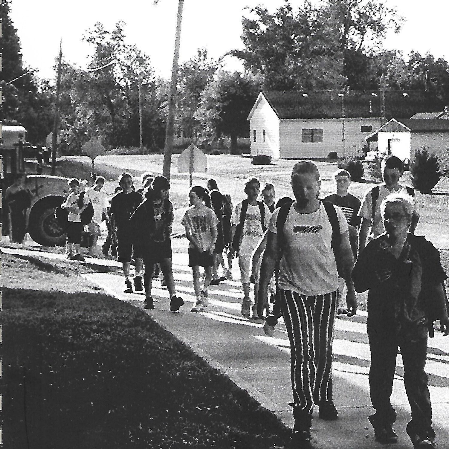 Black and white photo of a group of children walking on a sidewalk with a school bus on the street.