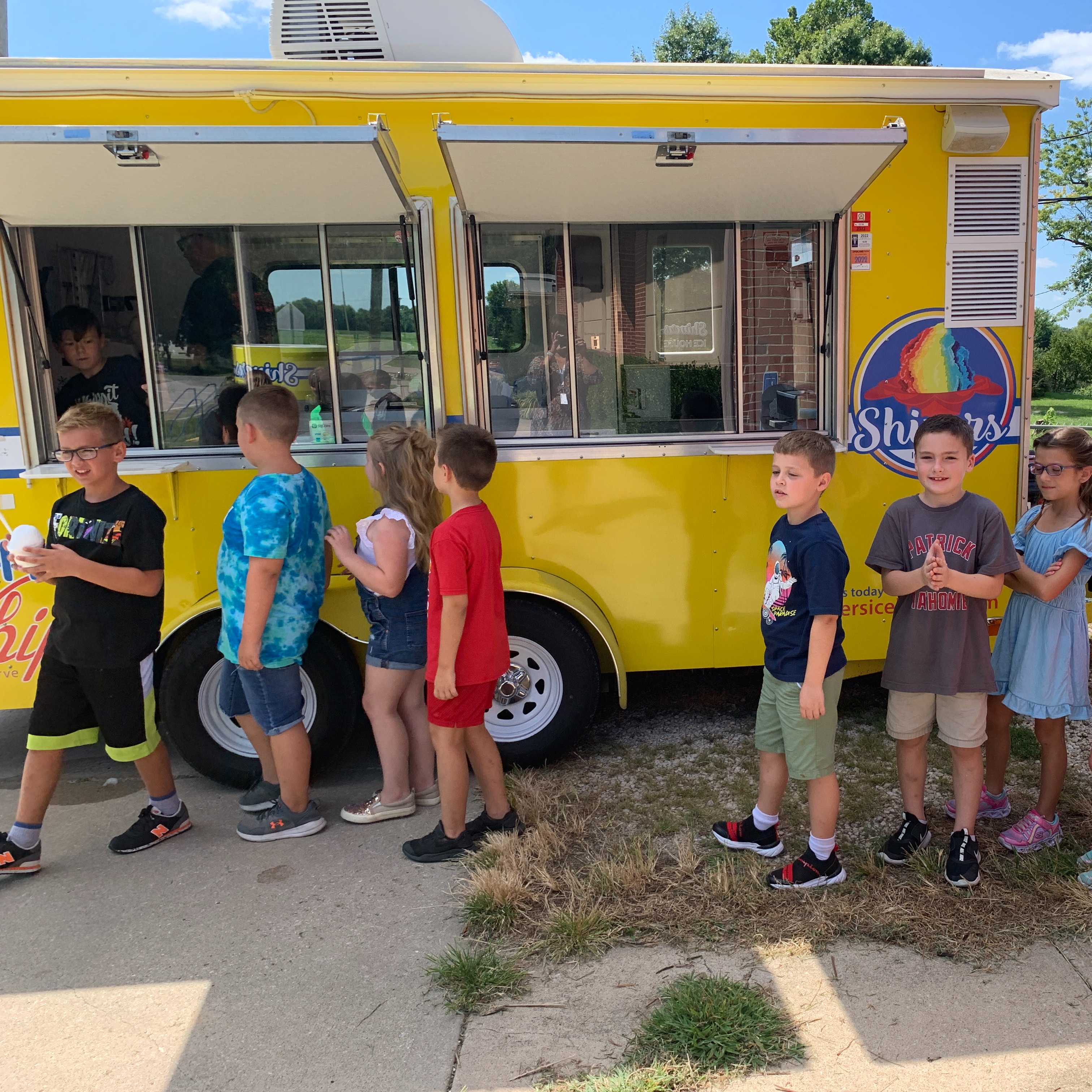 A line of children stand in front of a yellow ice cream truck. They wear casual summer clothes.