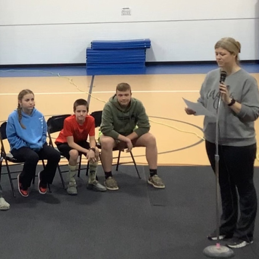 Group of people seated in chairs, facing a person standing on a mat, holding a paper.