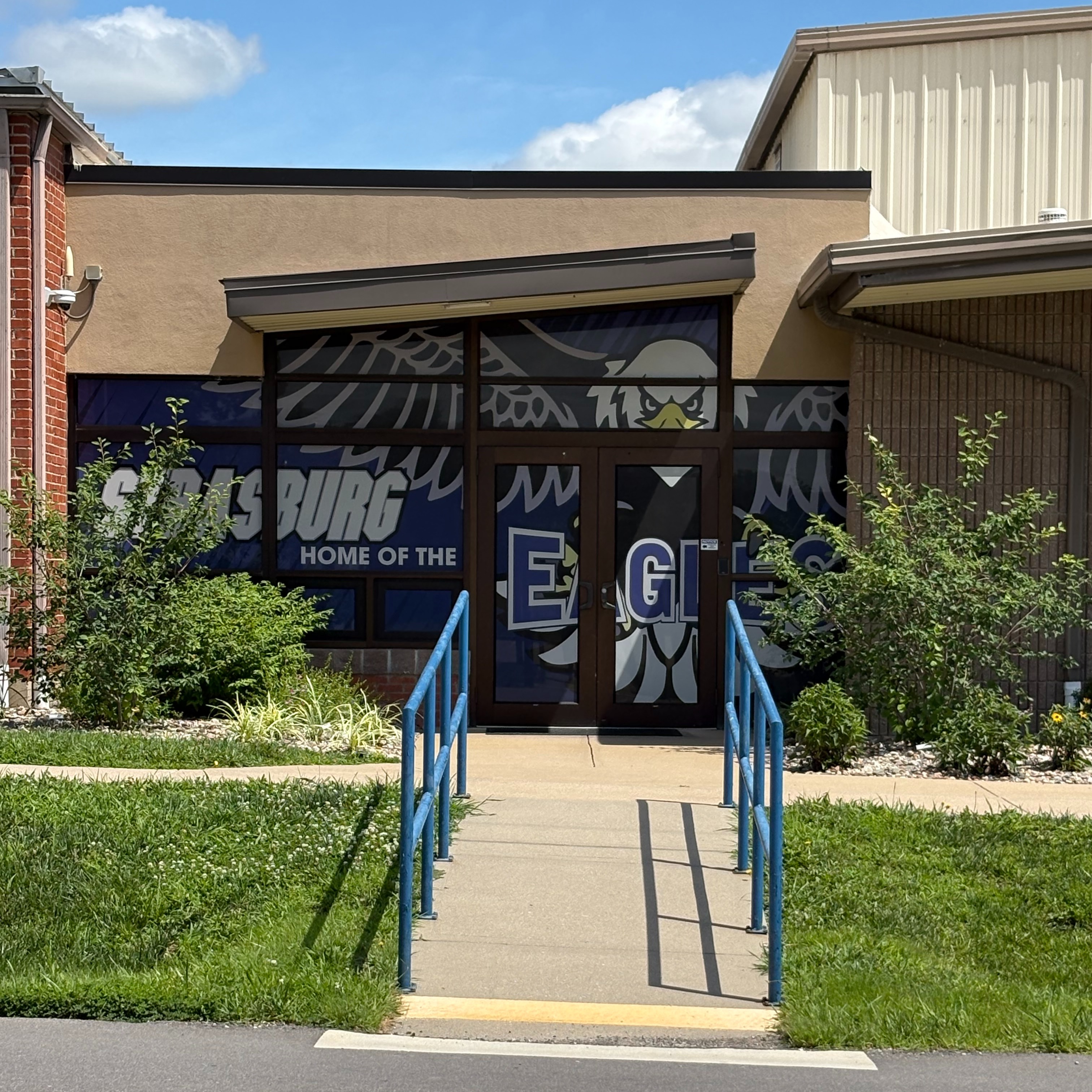 A building entrance with a ramp and blue railings. A blue and white sign on the entrance. Grassy lawn and shrubs.