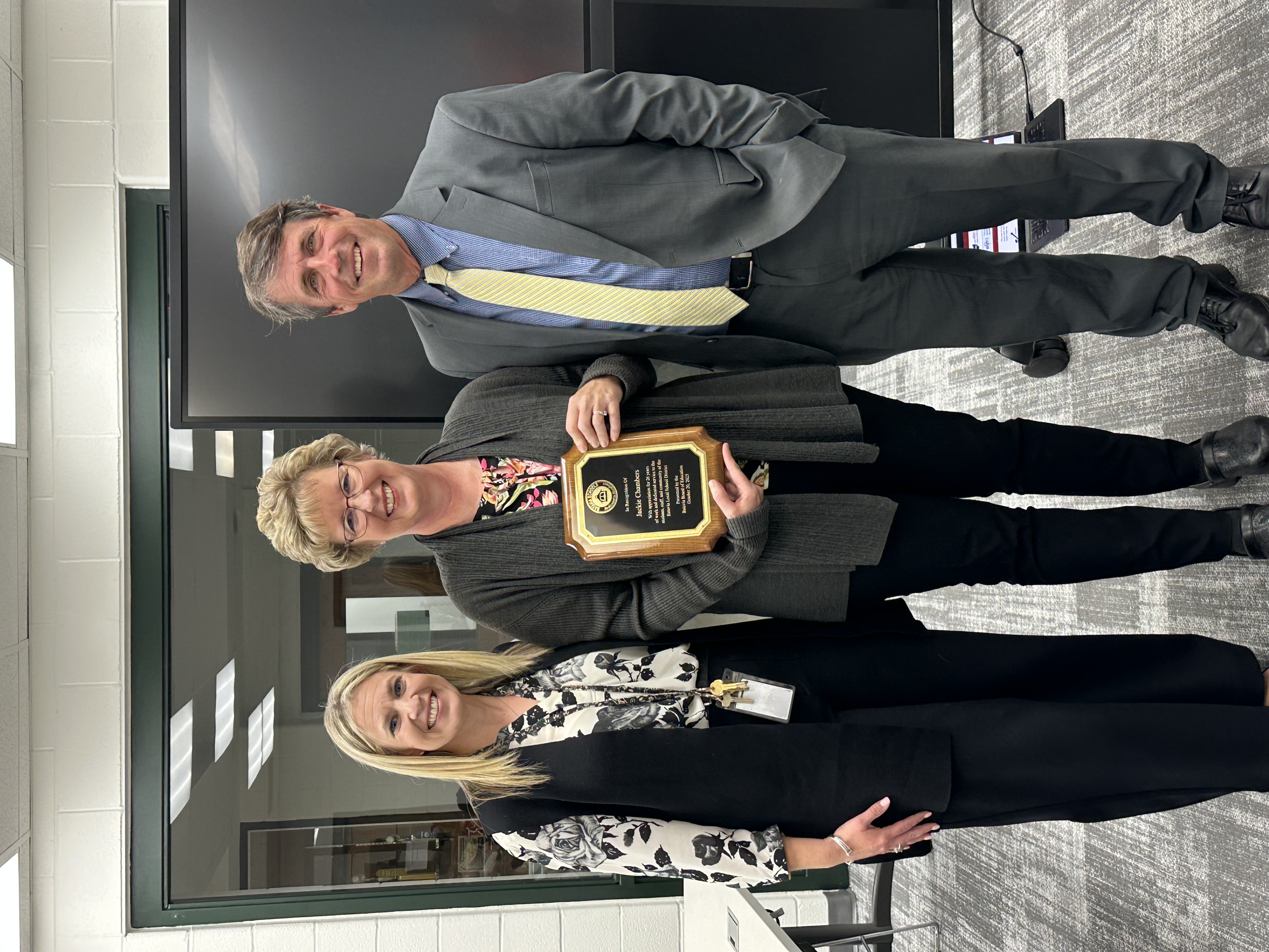 Image of staff member Jackie Chambers holding a plaque being recognized by principal liz Wainscott and superintendent Keith Millard