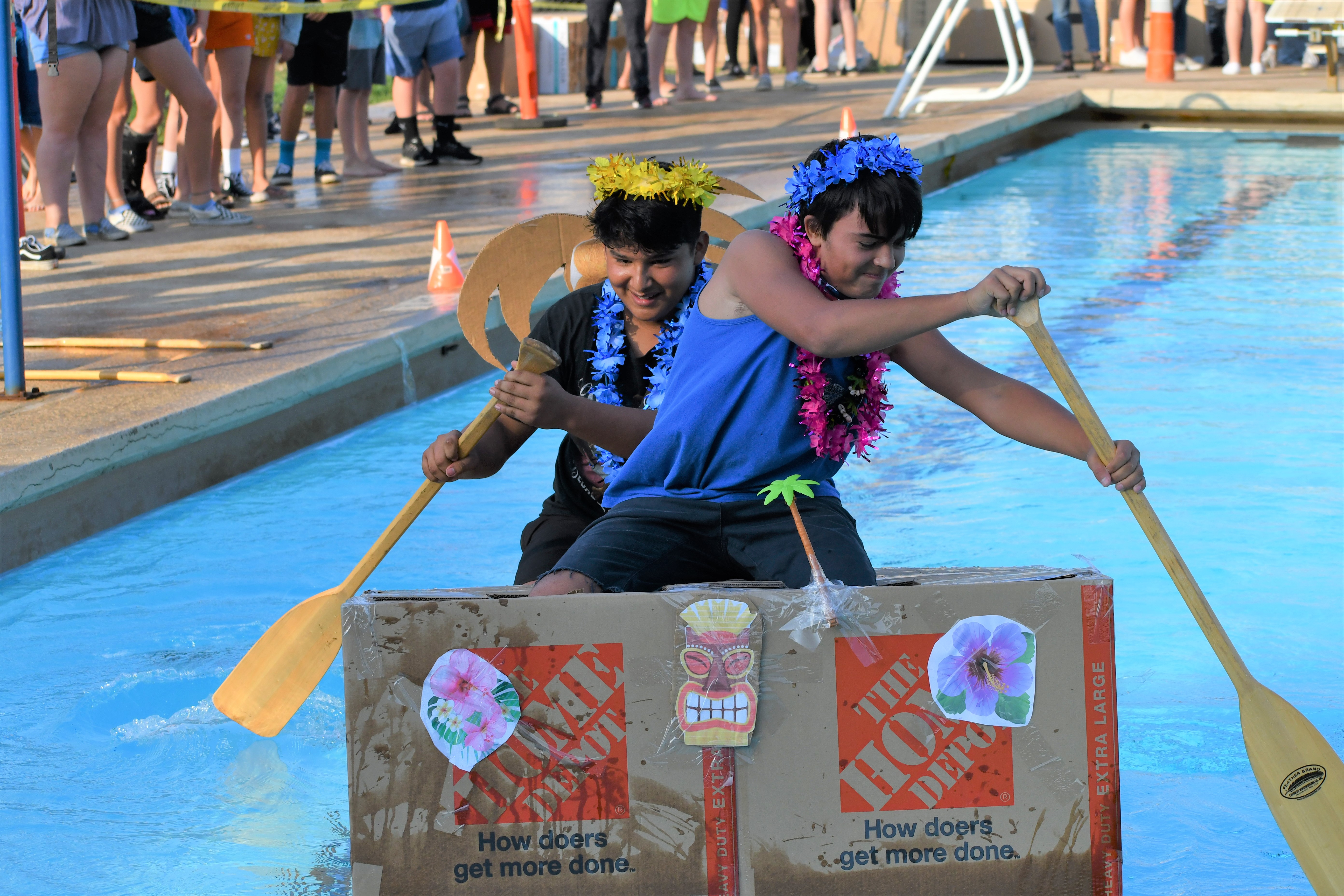 students rowing a cardboard box boat across a pool 