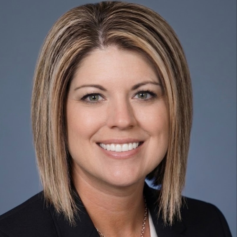 A woman with short blonde hair and a black blazer smiles in front of a blue backdrop