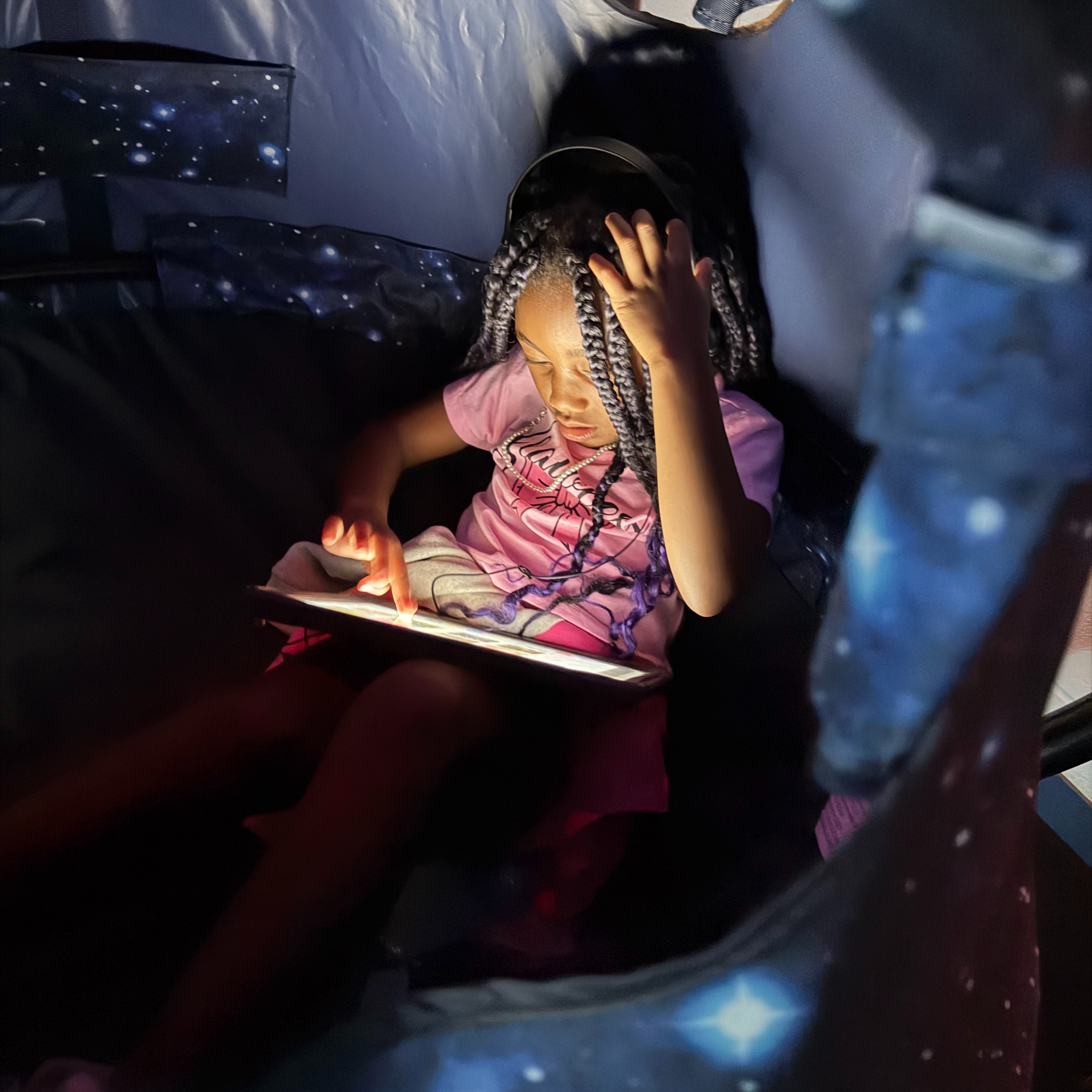 A child sits in a tent with a tablet, looking at the screen, and wearing headphones.