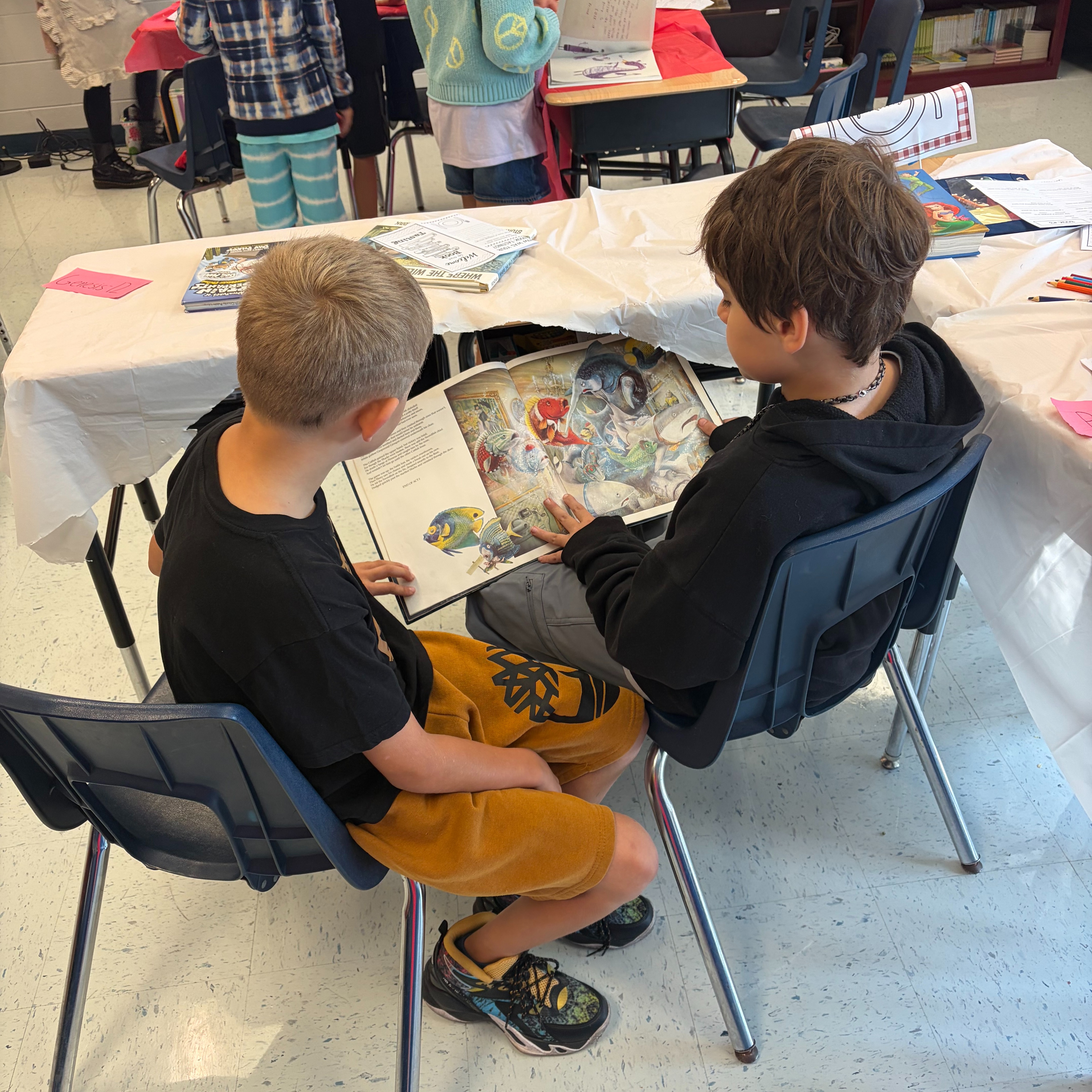 Two boys seated in chairs, one holding an open book, reading together at a long table.