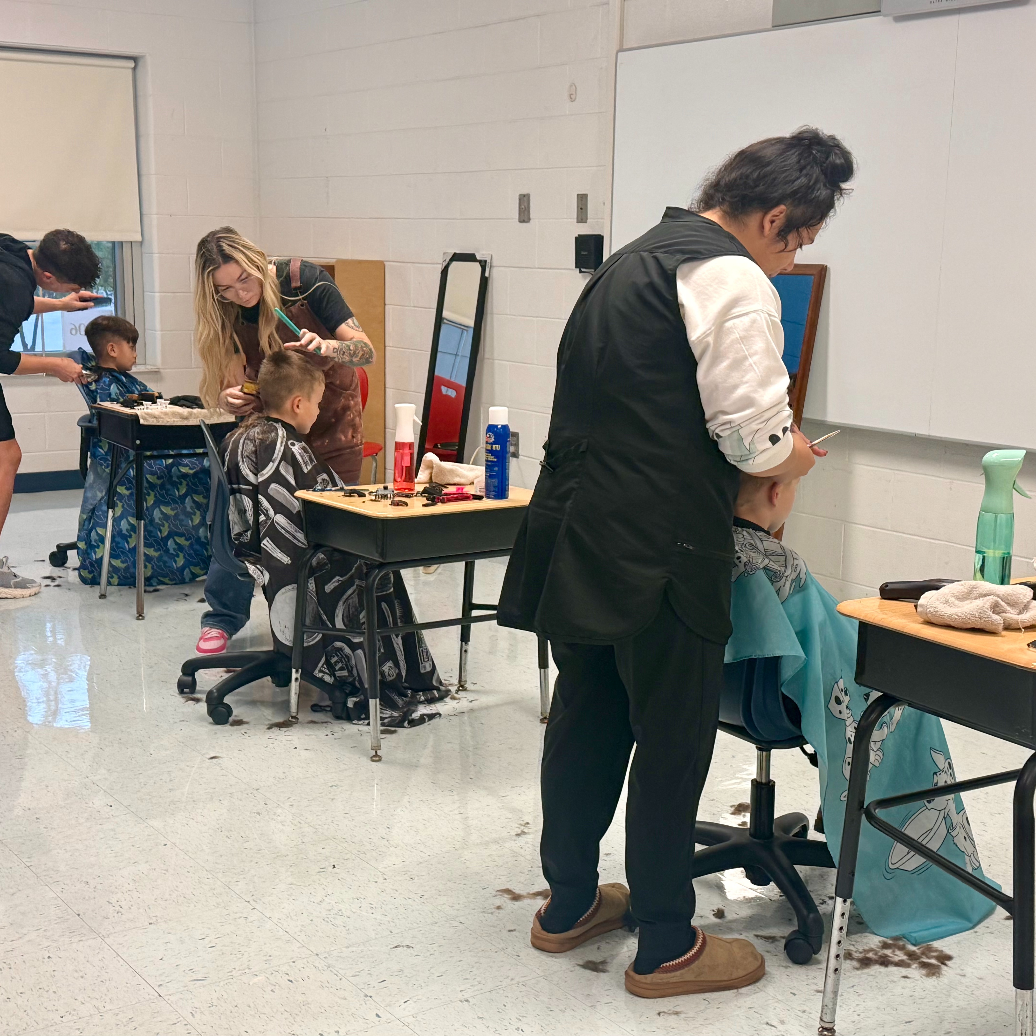 People getting their hair cut in a classroom. Some people have chairs with capes.
