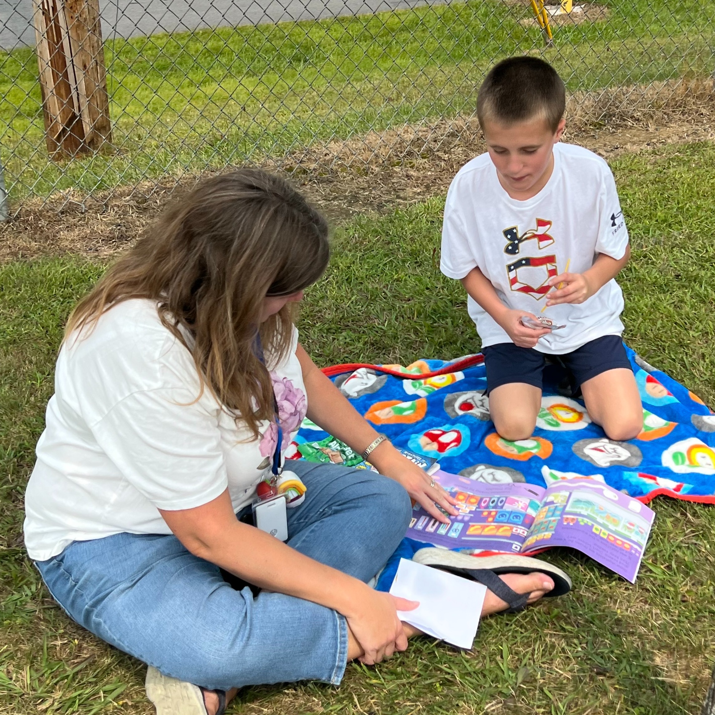 A woman and a child sit on a blanket on grass, reading a book. Behind them, a tree and a fence.