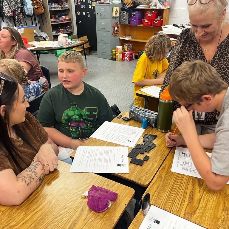 Several people sit at desks in a classroom, one with sunglasses on head. Three people look at papers.