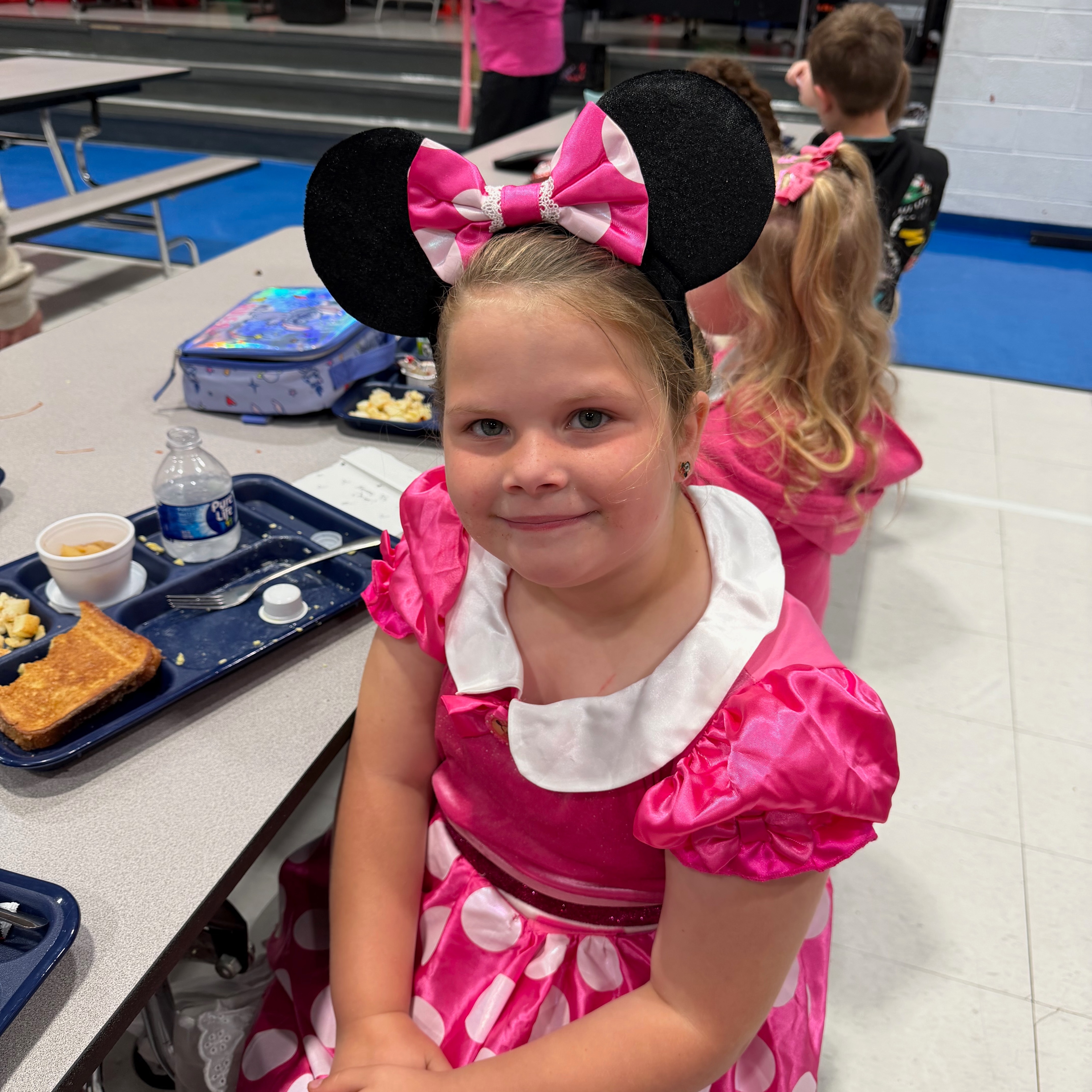 Child in pink Minnie Mouse dress sits at a lunch table, smiling, with lunch tray and others.